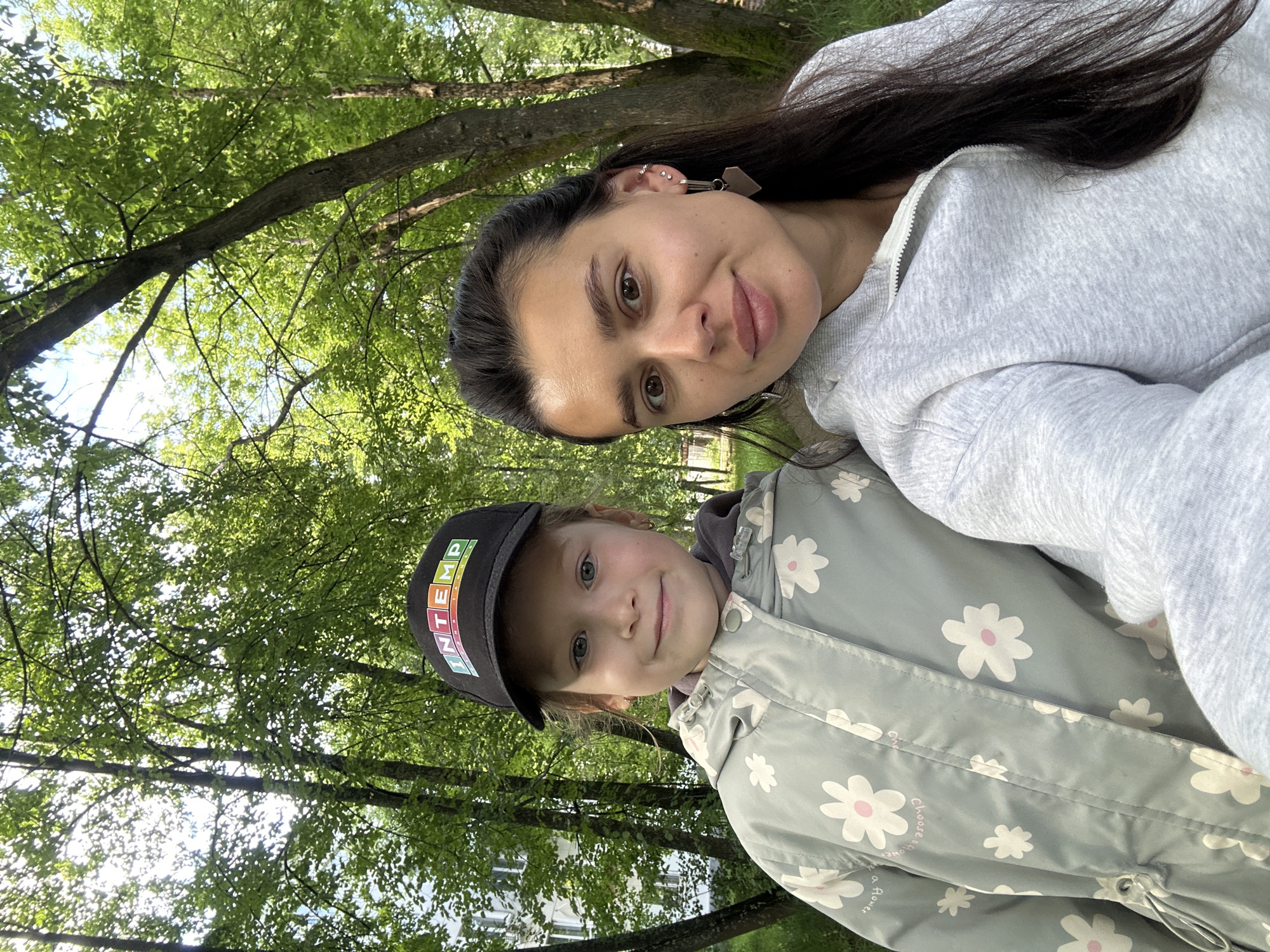 A woman and a young girl are posing for a selfie in an outdoor environment with trees and buildings in the background. The girl is wearing a black baseball cap with colorful letters that read "INTEMP" and a light gray jacket with a floral pattern. The woman is wearing a light gray hooded sweatshirt. The background shows lush green trees, sunlight filtering through the leaves, and a white building on the left. The setting appears to be a park or a residential area with greenery, possibly in the city of Chaikovsky, Russia, given the context. The overall scene depicts a casual, outdoor moment shared by the two individuals.