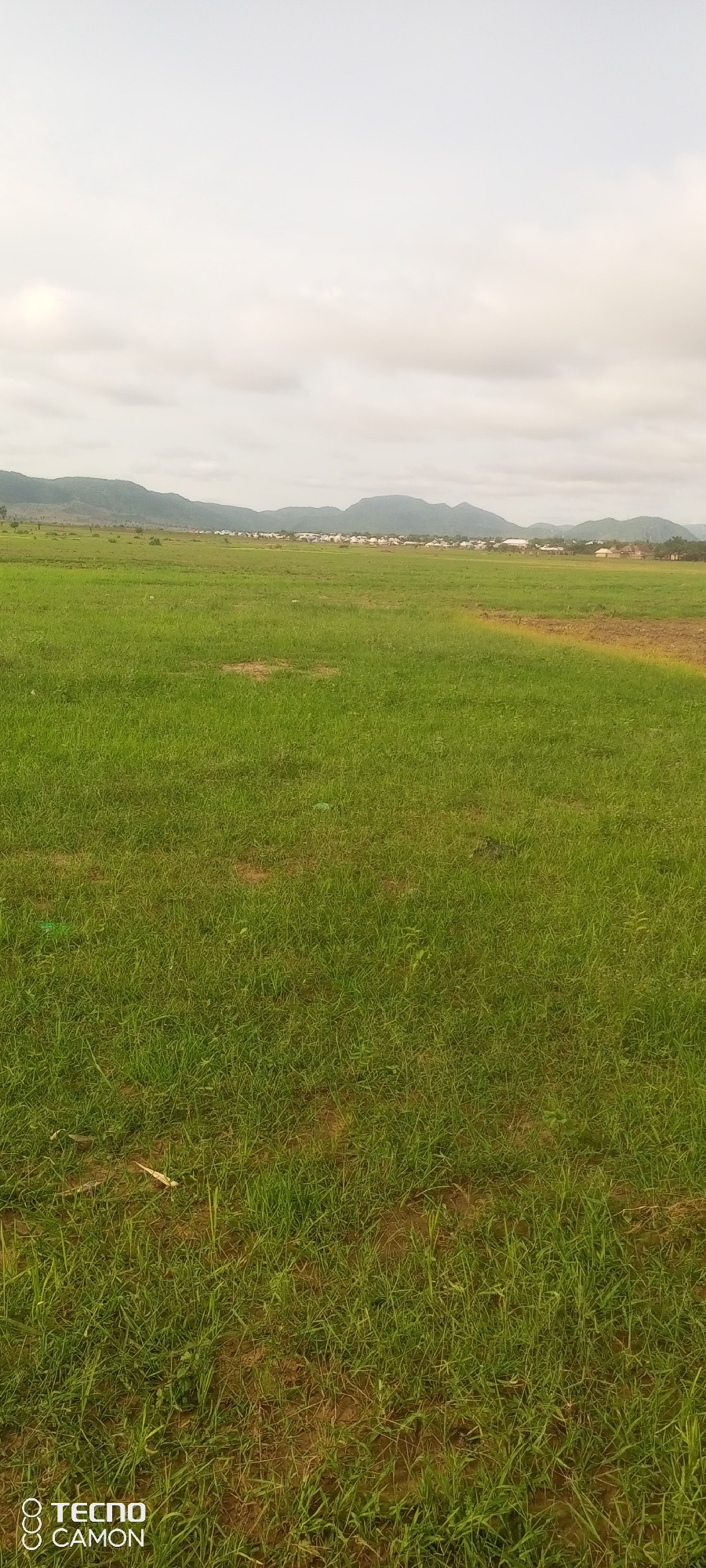 This is a vertical, eye-level outdoor photograph taken in Jalingo, Nigeria. The scene is dominated by a vast expanse of lush green grass, appearing well-maintained and possibly part of a field or park. In the distance, a line of buildings, likely a town or village, stretches across the horizon, nestled at the base of rolling hills and mountains. The sky is overcast with soft, diffused clouds, suggesting a mild or possibly rainy day. The lighting is even and lacks harsh shadows, further indicating cloudy conditions. The overall atmosphere is tranquil and natural, with no people or immediate activity visible. The foreground consists of the grassy field, with some patches of exposed earth visible, suggesting the grass may not be uniformly dense everywhere. The distant buildings are small and indistinct, providing a sense of scale to the landscape. The photograph also displays a watermark in the bottom left corner with the text "TECNO CAMON".