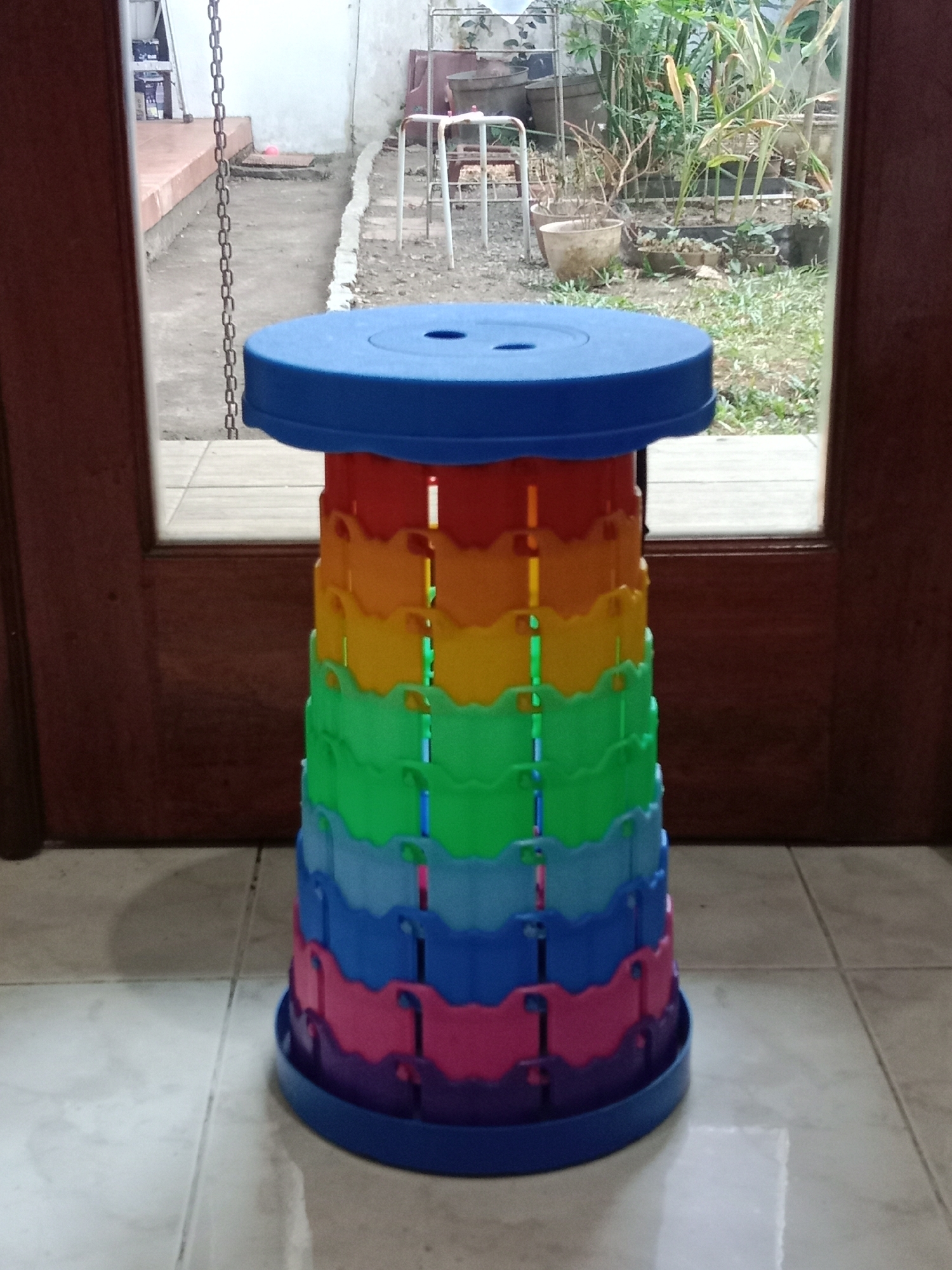 The image shows a colorful, collapsible stool in the foreground, positioned on a tiled floor. The stool is made of interlocking plastic segments in a rainbow of colors: blue, purple, pink, light blue, green, yellow, orange, and red, topped with a solid blue lid. Behind the stool, a doorway or large window reveals an outdoor area. Through the opening, we can see a paved walkway, some potted plants and vegetation, and the side of a building. The overall impression is of a domestic setting, possibly a backyard or patio. The description of the location as Ngingas, Indonesia, suggests a warm climate, consistent with the presence of lush plants. The image is taken from a medium distance, with the stool being the primary focus. There are no people or animals visible in the frame.