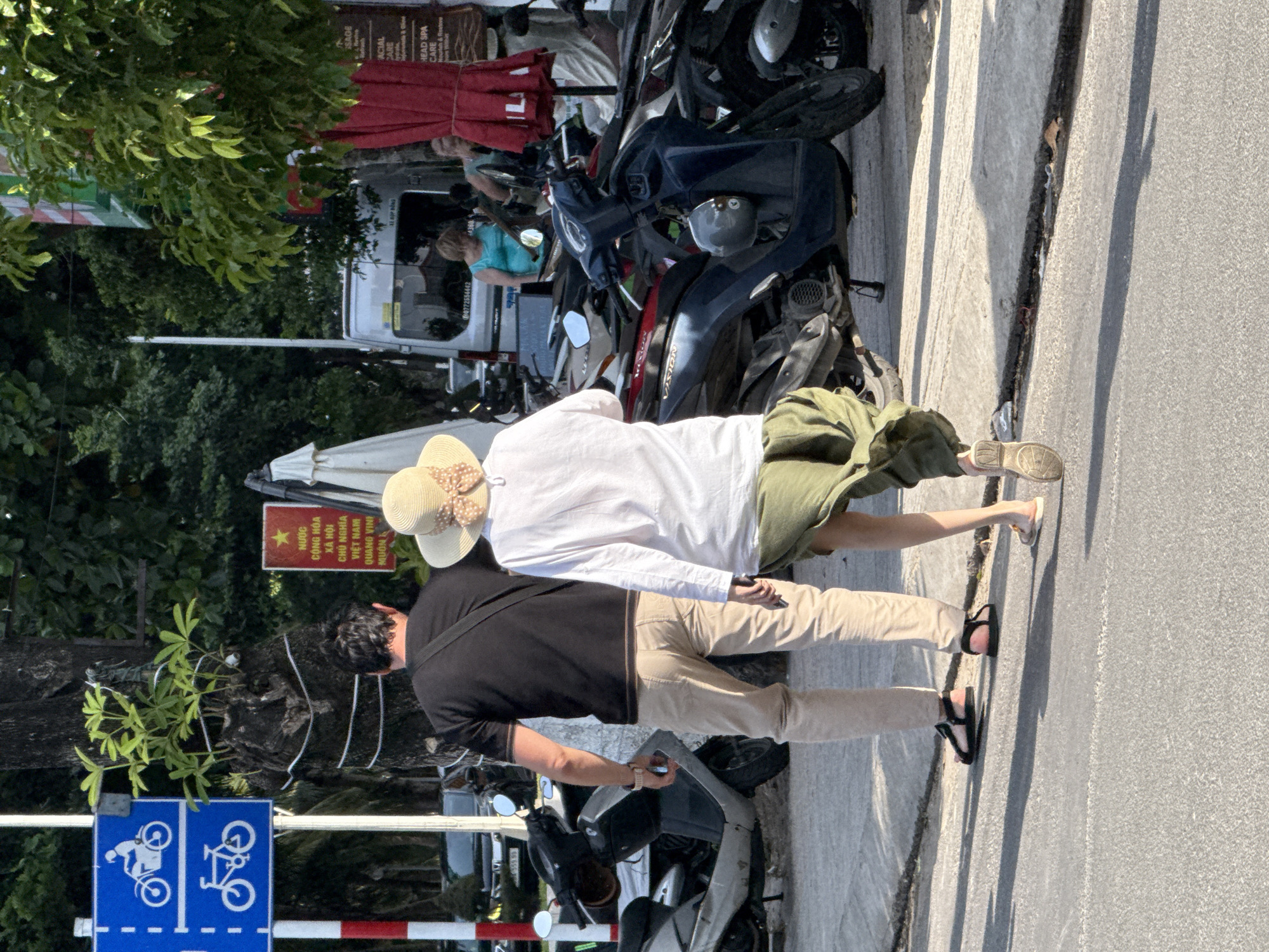 Two people, a man and a woman, walk away from the camera on a paved sidewalk in front of a row of parked motorbikes. The man, on the left, wears a black t-shirt and tan pants with black sandals. The woman, on the right, wears a white long-sleeved shirt, a wide-brimmed straw hat, and a flowing green skirt. She is holding a smartphone in her right hand. The motorbikes are parked along the sidewalk, with one blue scooter prominently in the foreground. In the background, there is a white van, trees, and buildings. A red sign with Vietnamese text and a star is visible on the left side of the image, near a tree trunk. A blue sign with white bicycle icons is also present on a pole to the left, indicating a cycling path. The scene appears to be a street in a city, possibly Bac Nha Trang Ward, Vietnam, given the presence of Vietnamese text on a sign and the abundance of motorbikes, which are common in Vietnam. The lighting suggests it is daytime, with shadows cast on the pavement.