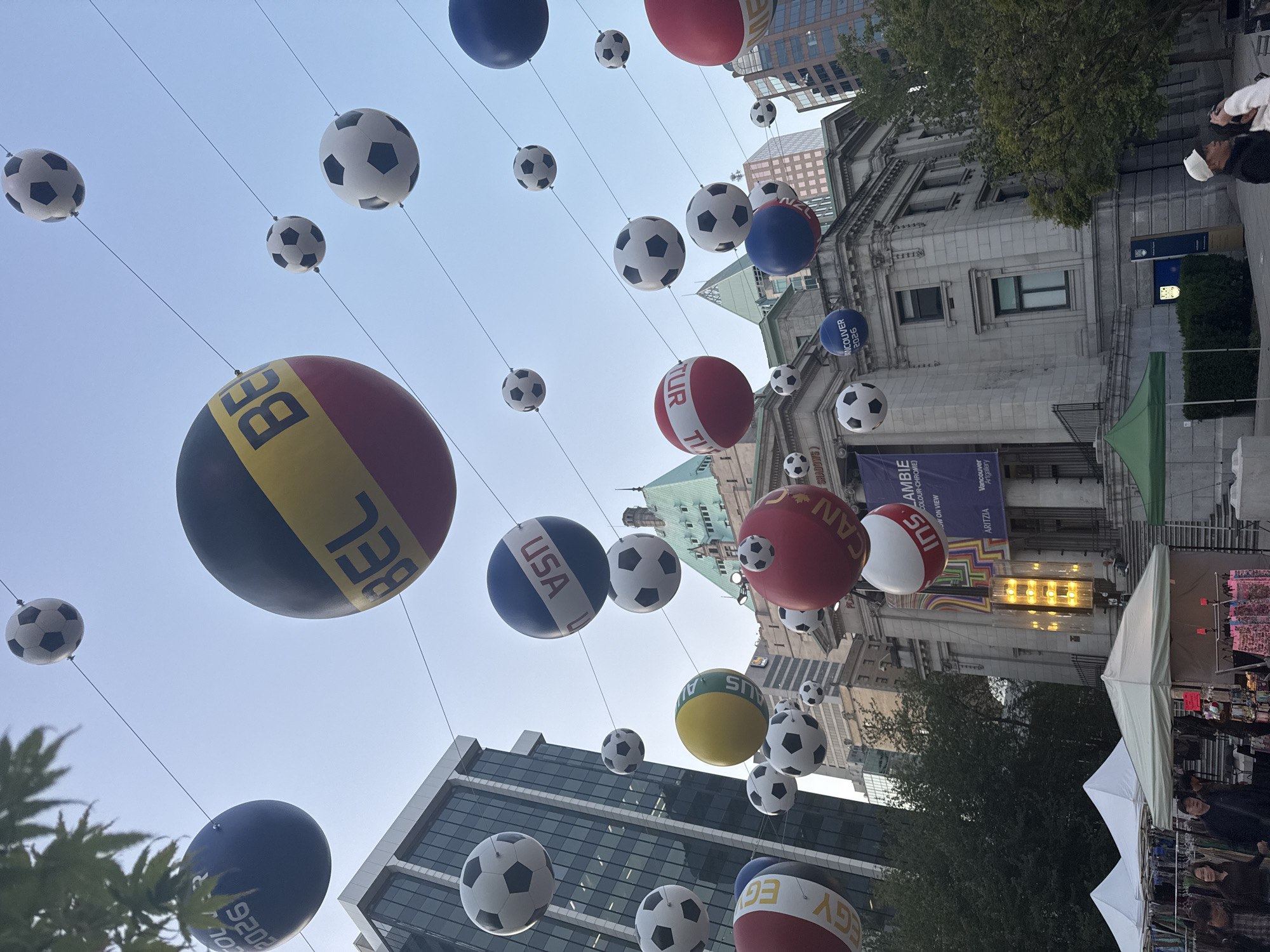 The image captures a street scene in Vancouver, Canada, featuring soccer balls with country abbreviations like "BEL," "USA," "CAN," "AUS," and "TUR" suspended in the air with thin wires. The background displays a mix of modern and historic buildings, including a large gray structure with a sign for the Vancouver Art Gallery. In the foreground, there are tents and market stalls with people browsing merchandise, indicating a street fair or market. The sky is light blue and hazy. The text “VANCOUVER 2026” is visible on the balloons.