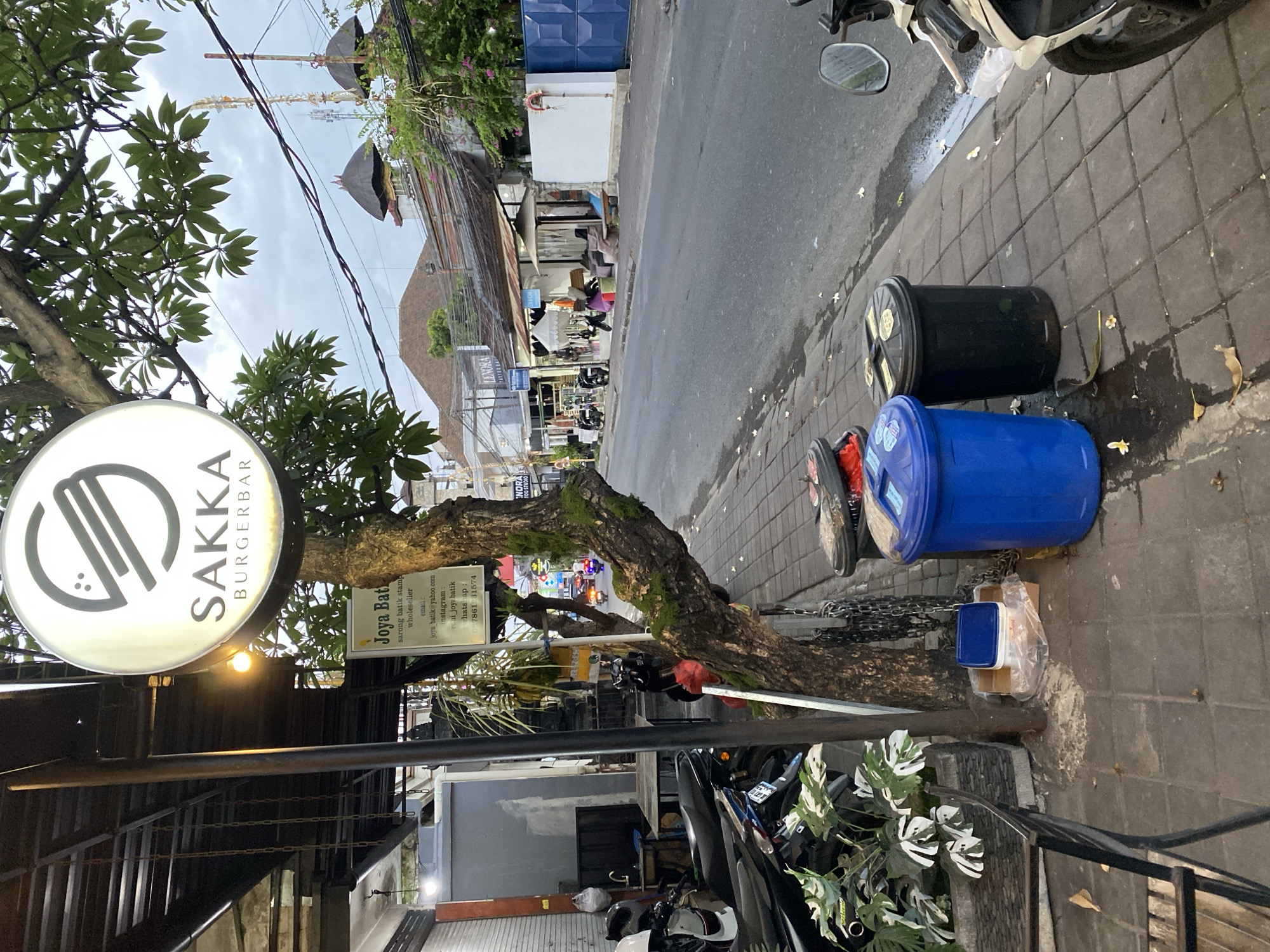 The image shows a street scene in North Kuta, Indonesia, with a focus on the sidewalk in front of a burger bar called "SAKKA BURGERBAR." A large, round sign with the restaurant's name and logo is prominently displayed on a tree. Several motorcycles are parked on the sidewalk, and two trash cans, one blue and one black, are situated near the restaurant. The street itself is paved and appears to be a typical urban road with buildings lining one side. The sky is overcast. A sign for "Joya Batik" is also visible, indicating other businesses in the vicinity. The overall impression is of a bustling commercial area with a mix of businesses and everyday life.