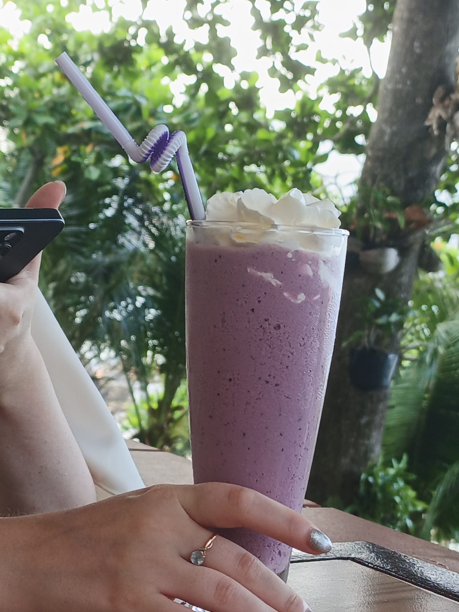 A close-up, waist-up shot shows a person holding a smartphone and a glass of blueberry smoothie. The person is wearing a ring with a light blue stone. The smoothie is topped with whipped cream and has a purple swirl straw. The background is blurred, showing lush green foliage and a tree trunk, suggesting an outdoor setting. The image is taken in Nha Trang, Vietnam.