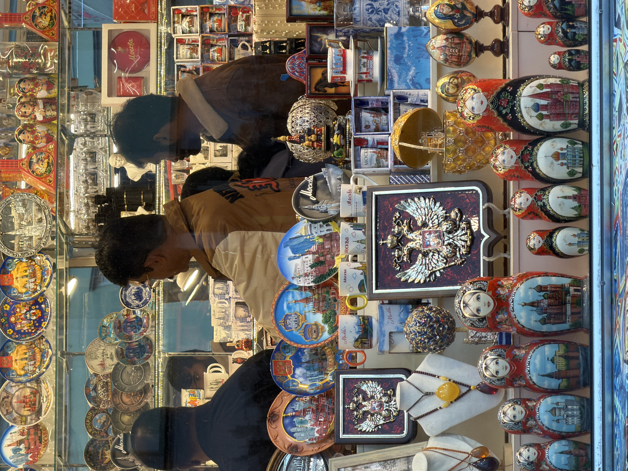 This image shows a display window of a souvenir shop in an unknown city. The window is filled with various Russian-themed souvenirs, including matryoshka dolls, decorative plates, cups, and painted eggs. In the background, two men are visible, appearing to be customers browsing the merchandise. The shop window is lit, illuminating the colorful items on display.