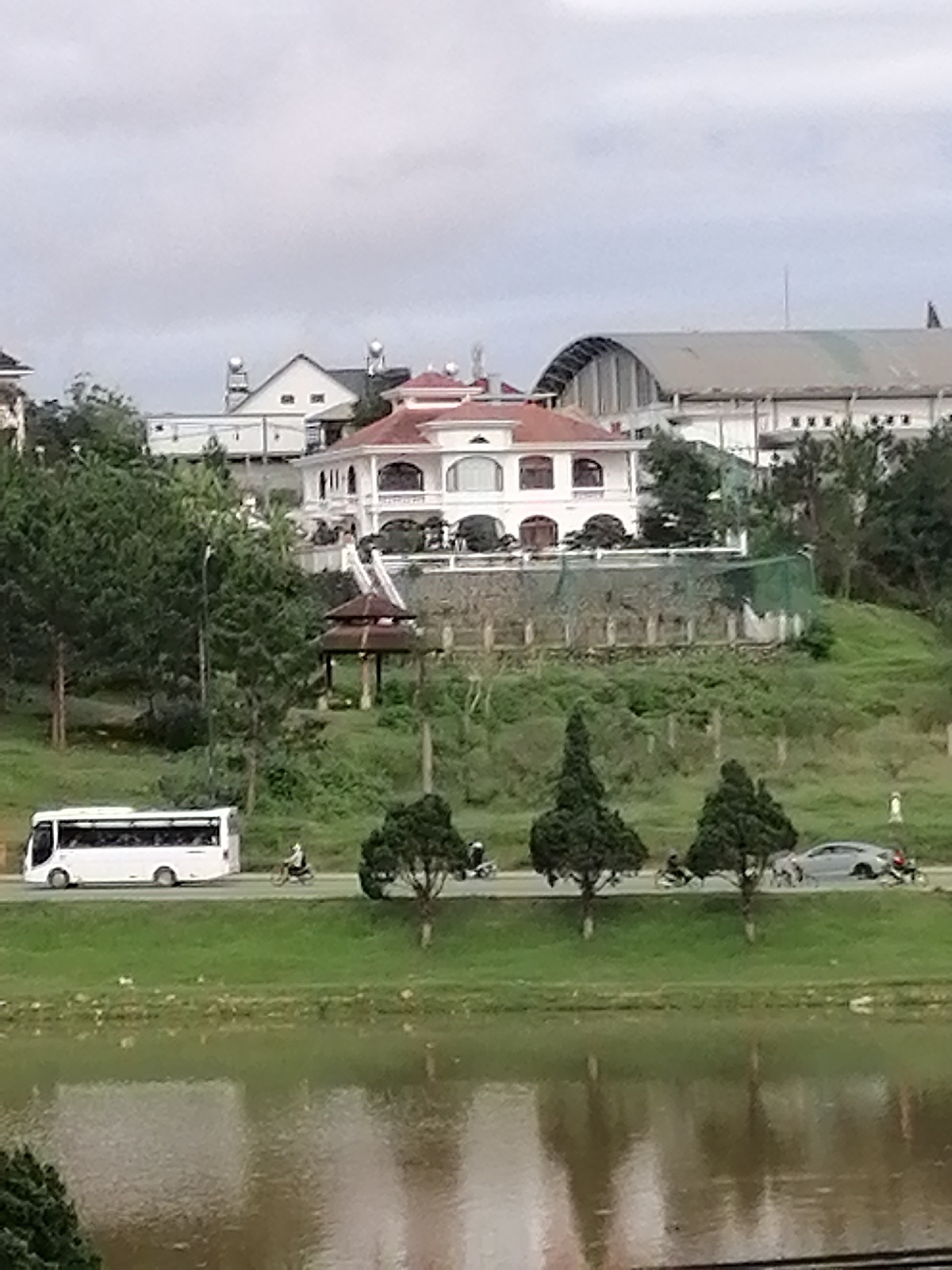 The image displays a panoramic view of a landscape in Da Lat, Vietnam, under an overcast sky. In the foreground, a calm body of water, likely a river or lake, reflects the muted light.
A vibrant green embankment separates the water from a paved road in the midground. Along this road, traffic is visible, including a white tour bus traveling left, a grey car, and several motorbikes with riders. A row of small, shapely evergreen trees lines the roadside, adding to the scenery.
Dominating the background is a grand, multi-story white building with a distinctive red-tiled roof, possibly a villa or mansion, situated on a lush green hill. The building showcases classical architectural elements with arched windows and balconies. Below it, on the slope, a traditional wooden gazebo with a tiered, pagoda-style roof adds a cultural accent. The hill is extensively covered with trees and dense greenery.
Further in the distance, behind the main villa, other structures are apparent: a white building with a gable roof, featuring two prominent round tanks or domes on its top, and a large industrial-style building with a sprawling, curved metallic roof, resembling an arena or large-scale facility.
The scene captures a blend of natural beauty, residential elegance, and functional infrastructure, typical of a developing urban area set within a verdant, hilly environment. The time of day is daytime, characterized by soft, diffused light due to the cloudy weather. No visible text is present in the image.