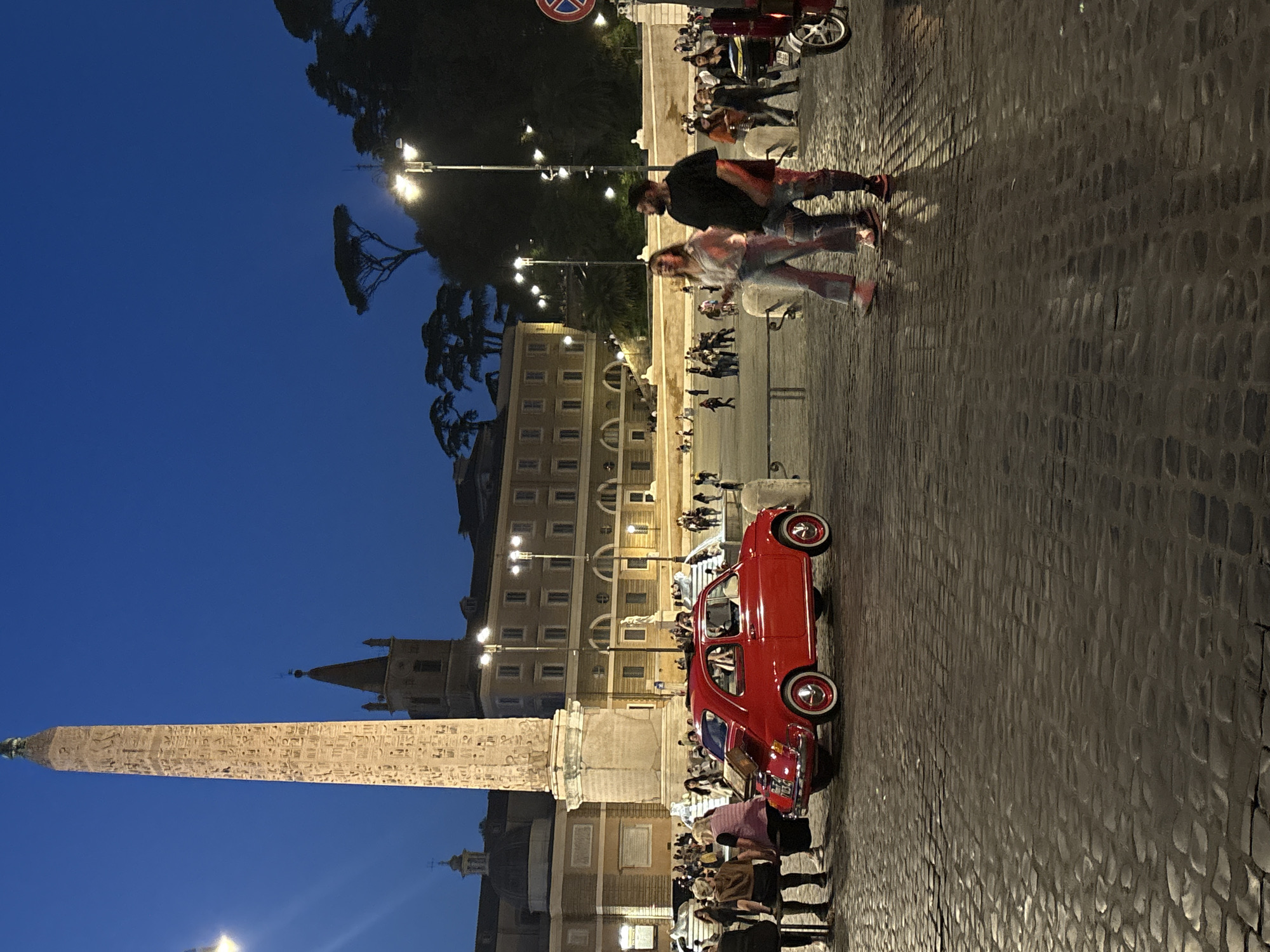 The image depicts Piazza del Popolo in Rome, Italy, during the evening. A tall obelisk stands prominently against a deep blue sky. Buildings are visible in the background, illuminated by streetlights. Numerous people populate the piazza. A red vintage car is parked on the cobblestone pavement. Several figures are walking, and others appear to be standing or gathered in groups.