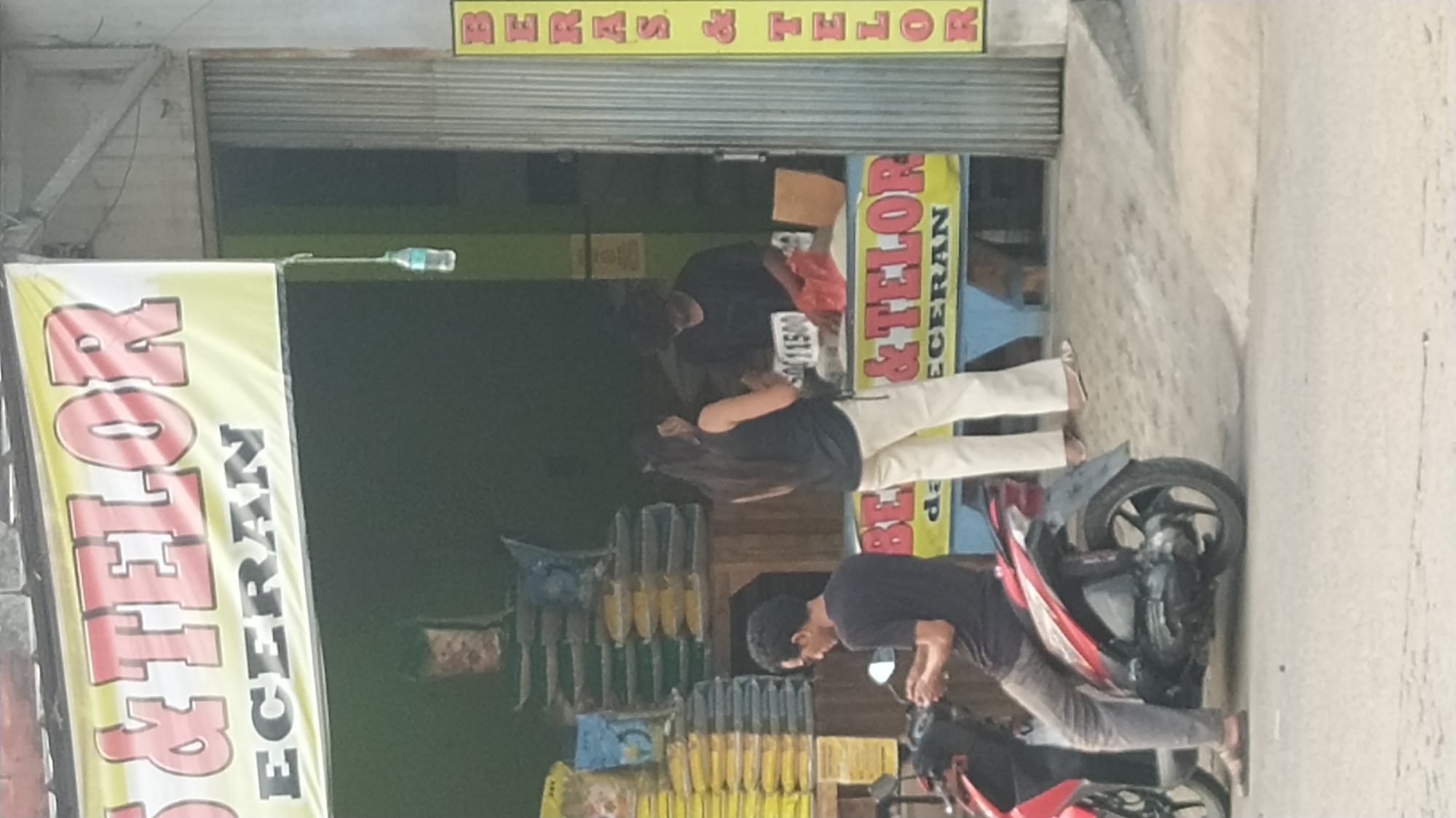 The image depicts an outdoor scene in front of a shop in Klapanunggal, Indonesia. A yellow banner with red and black lettering is prominently displayed, reading "BERAS & TELOR ECERAN" (Rice & Eggs Retail). To the right, a vertical yellow sign with red lettering reads "BERAS & TELOR".
In the foreground, a person is seated on a red and black motorcycle. They are wearing a dark t-shirt and grey pants, with their feet bare. The motorcycle is parked on a grey, textured surface that appears to be a road or pathway.
Further back, two people are interacting in front of a counter within the shop. One individual, wearing a black tank top and cream-colored pants, stands facing away from the camera. They appear to be interacting with another individual who is partially obscured by the counter. The second person, wearing a black t-shirt and dark pants, is leaning over the counter. A red plastic bag and a white object, possibly a price tag with the number "11500" visible, are on the counter.
Behind the counter, stacks of bagged goods, likely rice, are visible, with different colored bags. The shop itself has a green interior. A plastic water bottle is hanging from a string outside the shop. The overall environment suggests a commercial area, possibly a small grocery store or market stall.