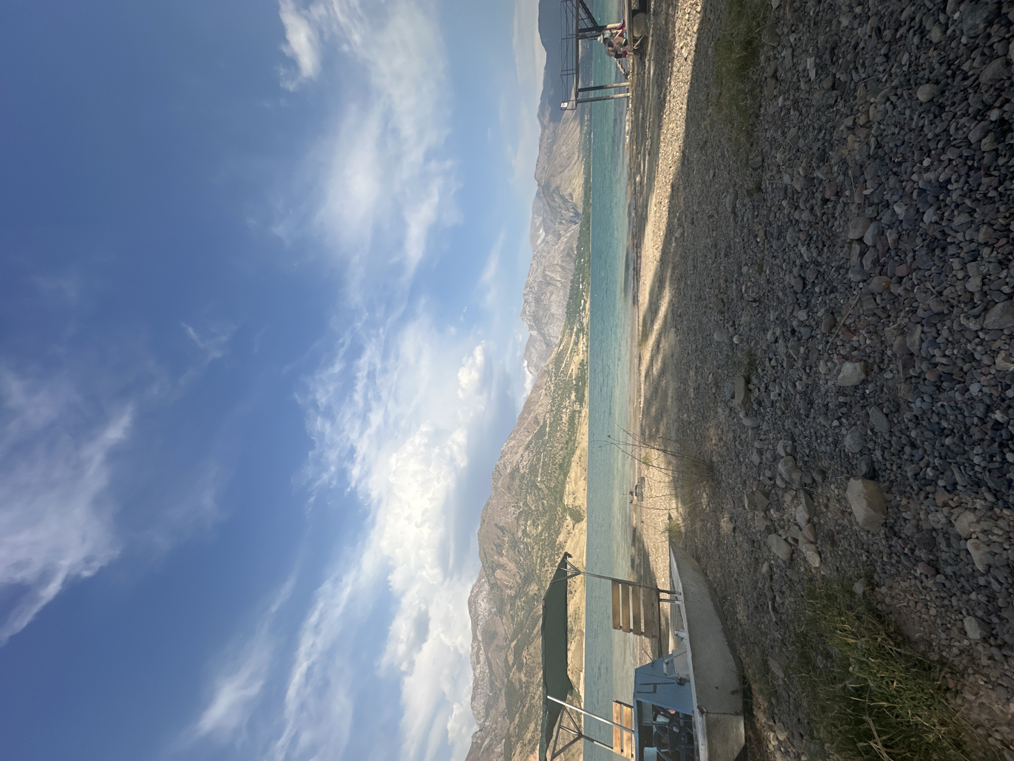 The image depicts a scenic view of a rocky shore leading to a body of water, with a mountainous landscape in the background. The sky is partly cloudy with a bright blue hue. On the left side of the shore, a small boat with a green canopy and wooden seats is beached. The water appears calm with gentle ripples. On the right side, a wooden pier extends into the water, with two individuals seated on its edge. The environment suggests a tranquil outdoor setting, likely a reservoir or lake, given the mountainous terrain. The location is identified as Yusuphana, Uzbekistan.