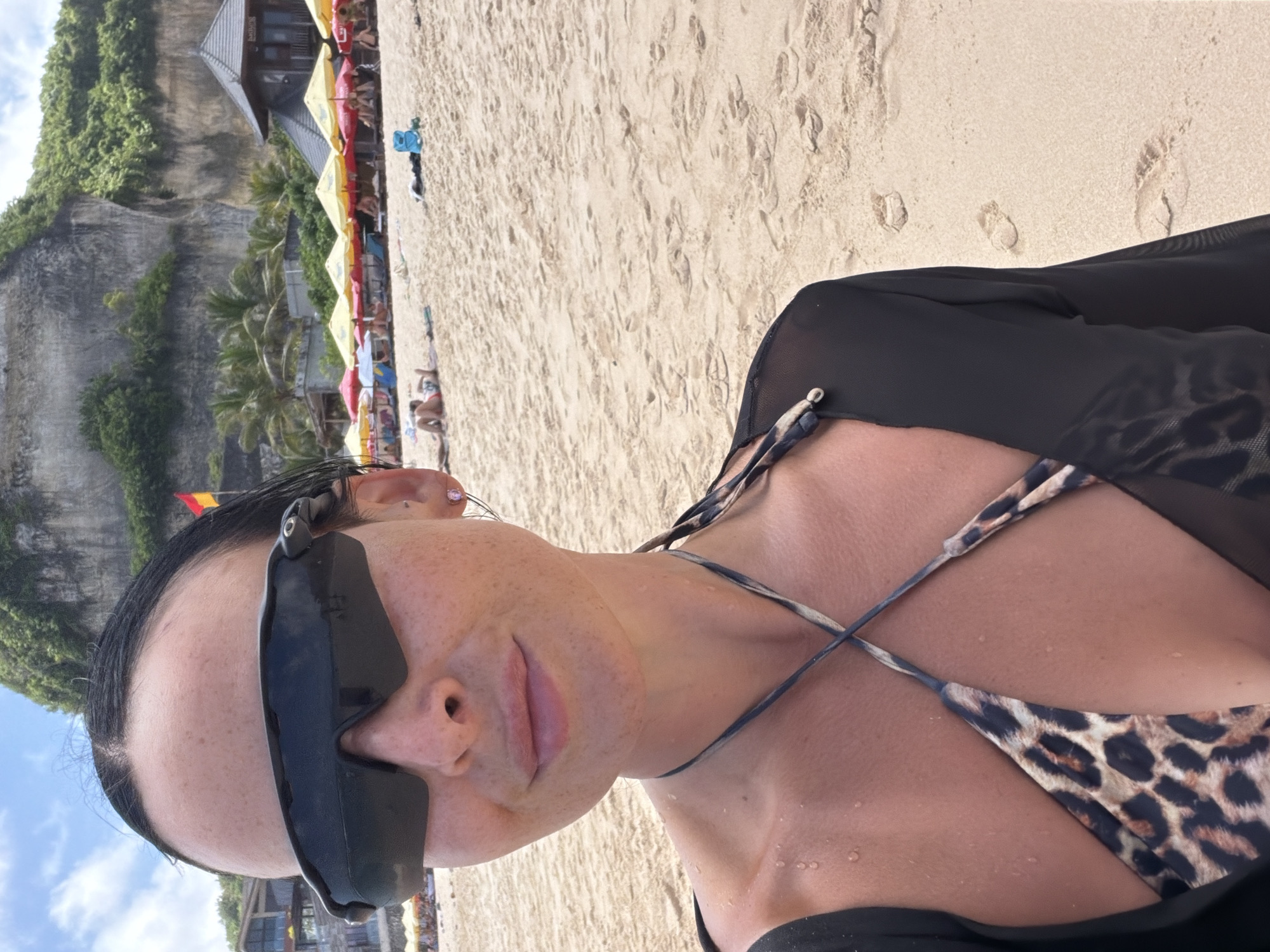 A waist-up, canted angle close-up photo depicts a woman with sunglasses in a leopard print bikini and black cover-up on a beach. The location is Padang Padang Beach in Ungasan, Indonesia. Behind her are beachgoers, colorful umbrellas, restaurants, and rock cliffs with trees. The woman is looking toward the camera. The beach sand has footprints.