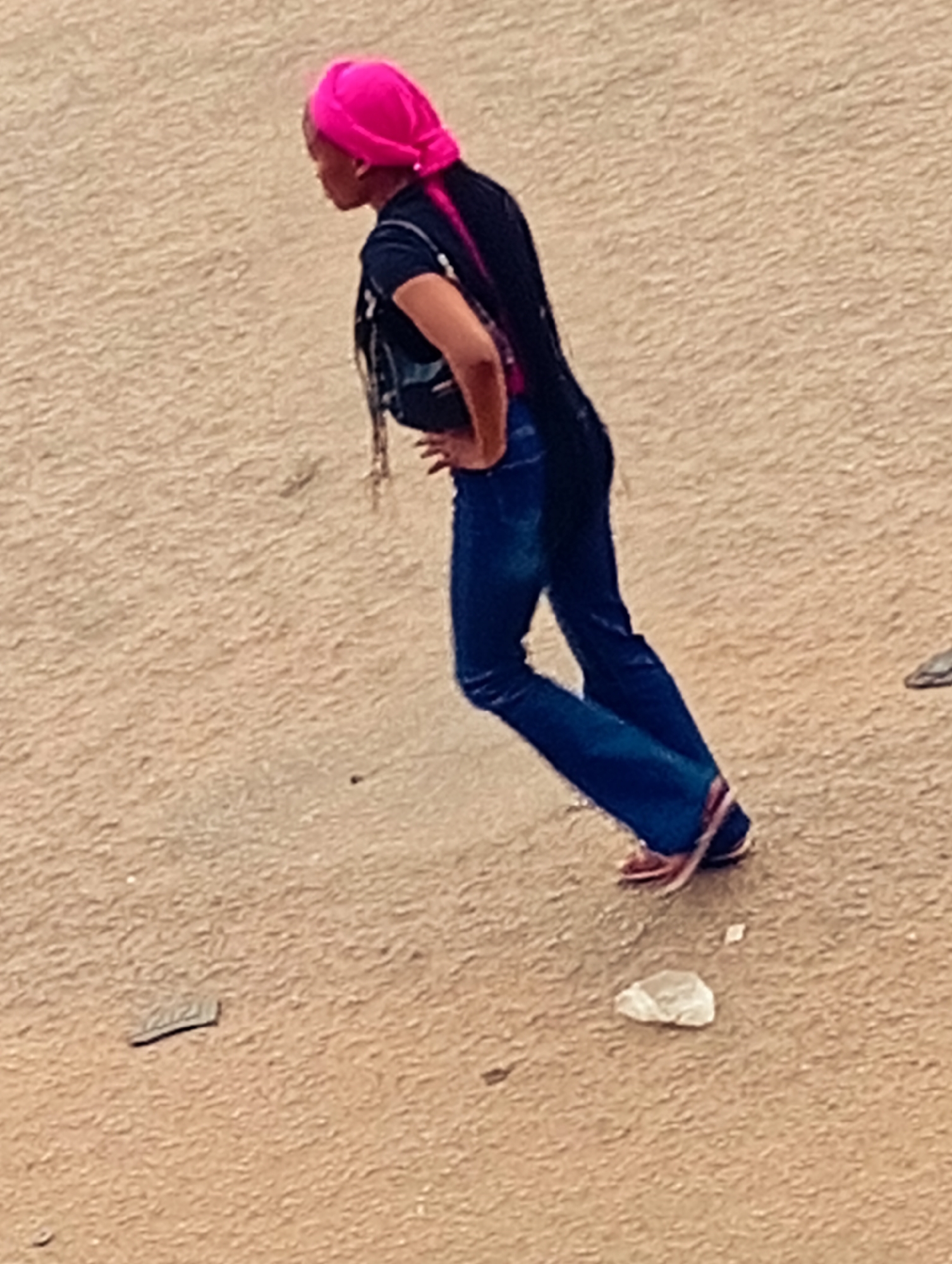 A woman wearing a bright pink head covering, a black top, blue jeans, and sandals is walking on a dirt or sand surface. She has a small black bag slung over her shoulder. The location is Bukuru, Nigeria. There is scattered debris on the ground, including what appears to be a piece of paper or plastic. The woman is positioned at a slight angle, appearing to walk away from the camera's perspective.