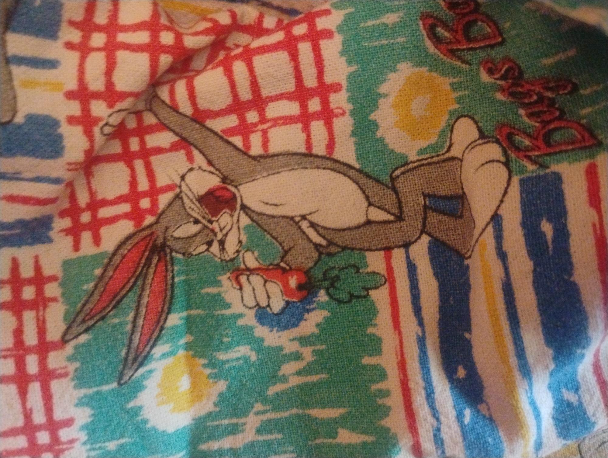 The image displays a close-up of a textile material, possibly a piece of clothing or fabric, featuring a cartoon character. The character is Bugs Bunny, depicted in his typical gray fur with long ears, white belly, and buck teeth, mouth open in a wide smile. He is holding a red object, possibly a carrot or a toy, in his left hand. The background is a busy pattern of abstract shapes and lines in various colors, including teal, green, yellow, red, and white. There are also some red and white plaid elements. In the lower right corner, text in a stylized red font reads "Bags B". The fabric appears to be a textured weave. There is no indication of a specific location in the city of Rumyantsevo, Bulgaria, nor any context that would suggest it.