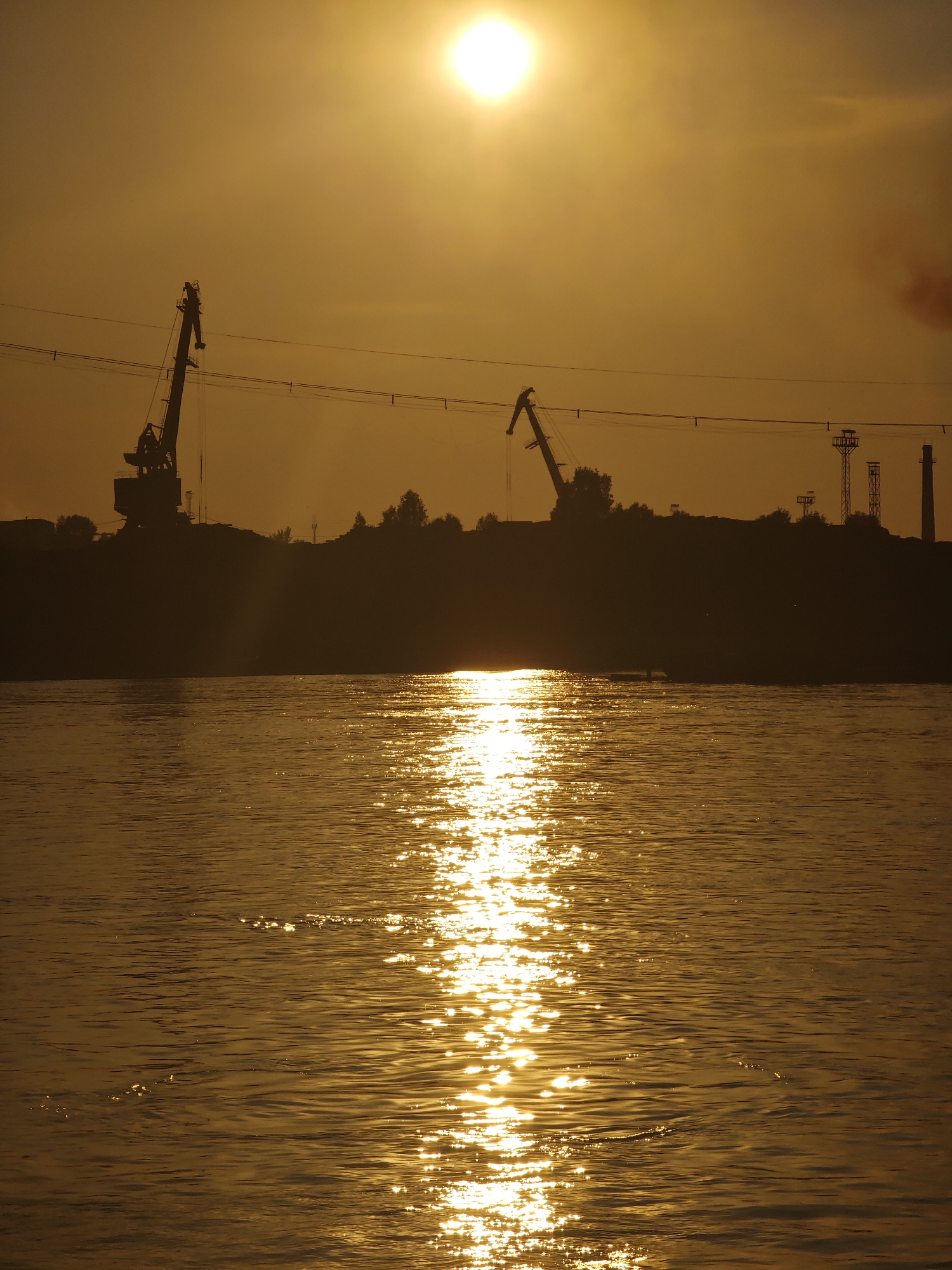 This is a photograph of a sunset over a body of water, likely a river, in the city of Lesosibirsk, Russia. The sun, a bright orb in the upper center of the frame, casts a warm, golden light across the scene. The water in the foreground reflects the sunlight, creating a shimmering path of light that stretches across the surface. In the background, silhouetted against the bright sky, are several industrial cranes and structures, suggesting a port or industrial area. Trees and other vegetation are visible along the distant shore, forming a dark line against the horizon. The overall atmosphere is serene and majestic, capturing the beauty of a natural phenomenon in an industrial setting.