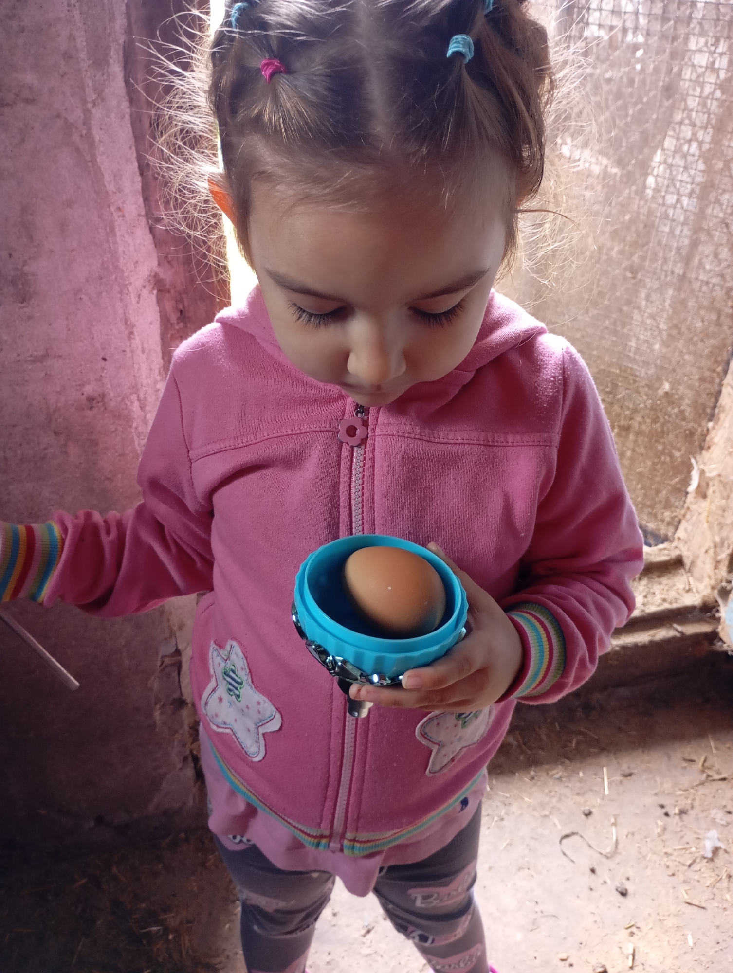 A young girl with dark hair tied in small pigtails with pink and blue bands is holding a brown egg in a small blue container. She is wearing a pink hooded jacket with patches of a white star with a pink outline and a smiling face on it, and a pink stripe with green and yellow stripes around the cuffs. Her grey leggings have a white and pink striped pattern. She is looking down at the egg. The background appears to be an interior with rough, pinkish-colored walls, and a loosely woven material or netting in the upper right corner. The floor is covered with dirt and straw. The lighting suggests an indoor environment, possibly a rural or farm setting. The image does not provide specific information that would allow for a precise location within Horishni Plavni, Ukraine, or any identifiable city landmarks.