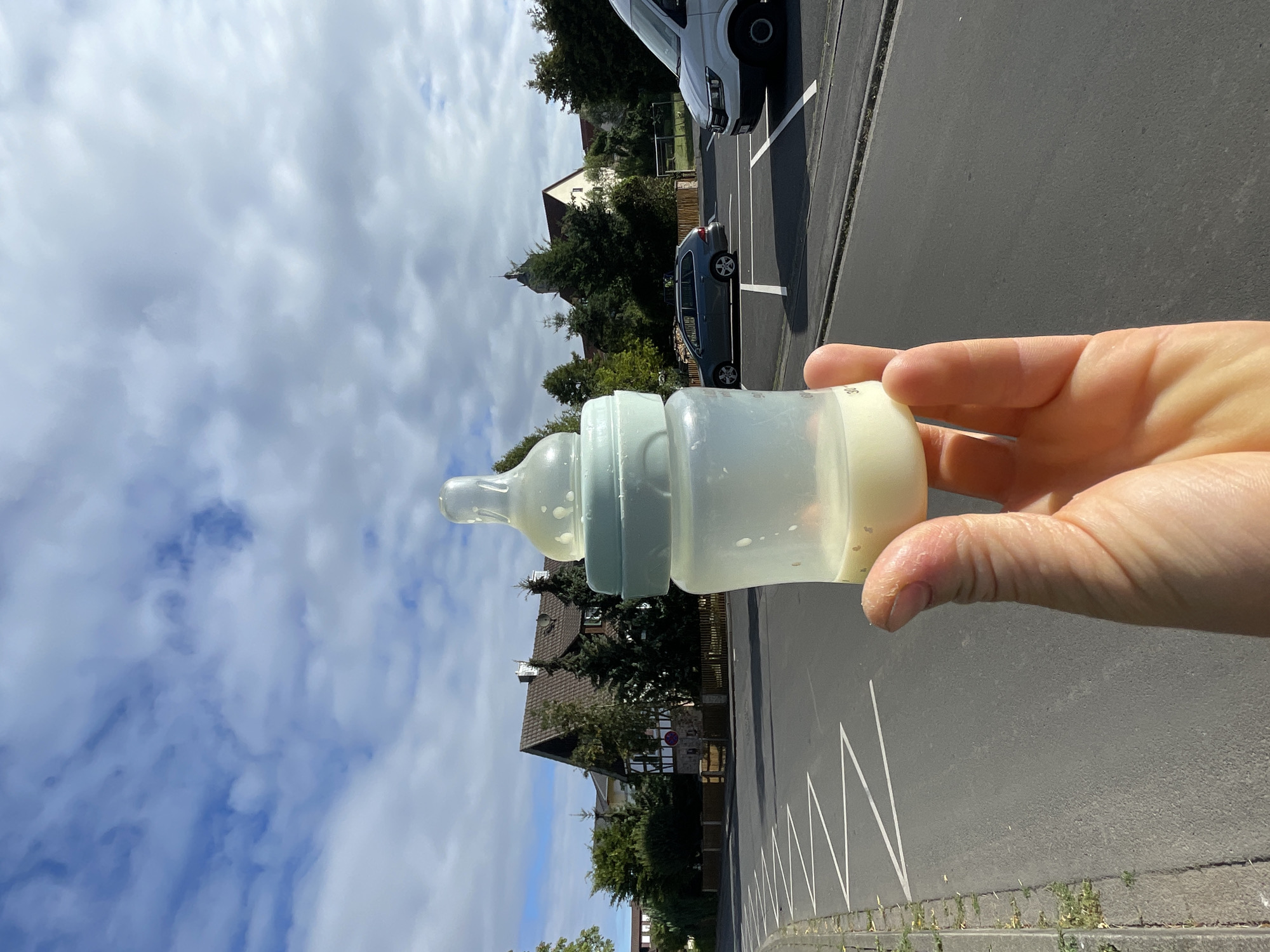 An out-of-focus, close-up shot features a human hand holding a baby bottle filled with a light-colored liquid. The bottle, made of clear plastic with a pale blue cap and nipple, is positioned in the foreground, obscuring the background. Beyond the bottle, a street is visible, lined with parked cars and houses with traditional German architecture, suggesting a residential area. A white van is parked on the right side of the frame, and a gray station wagon is visible on the left. The sky is blue with scattered clouds. The presence of houses, parked cars, and a street indicate an urban or suburban setting. The architecture of the houses, specifically the timber-framed elements on the house in the middle background, are characteristic of Neukirchen, Germany.