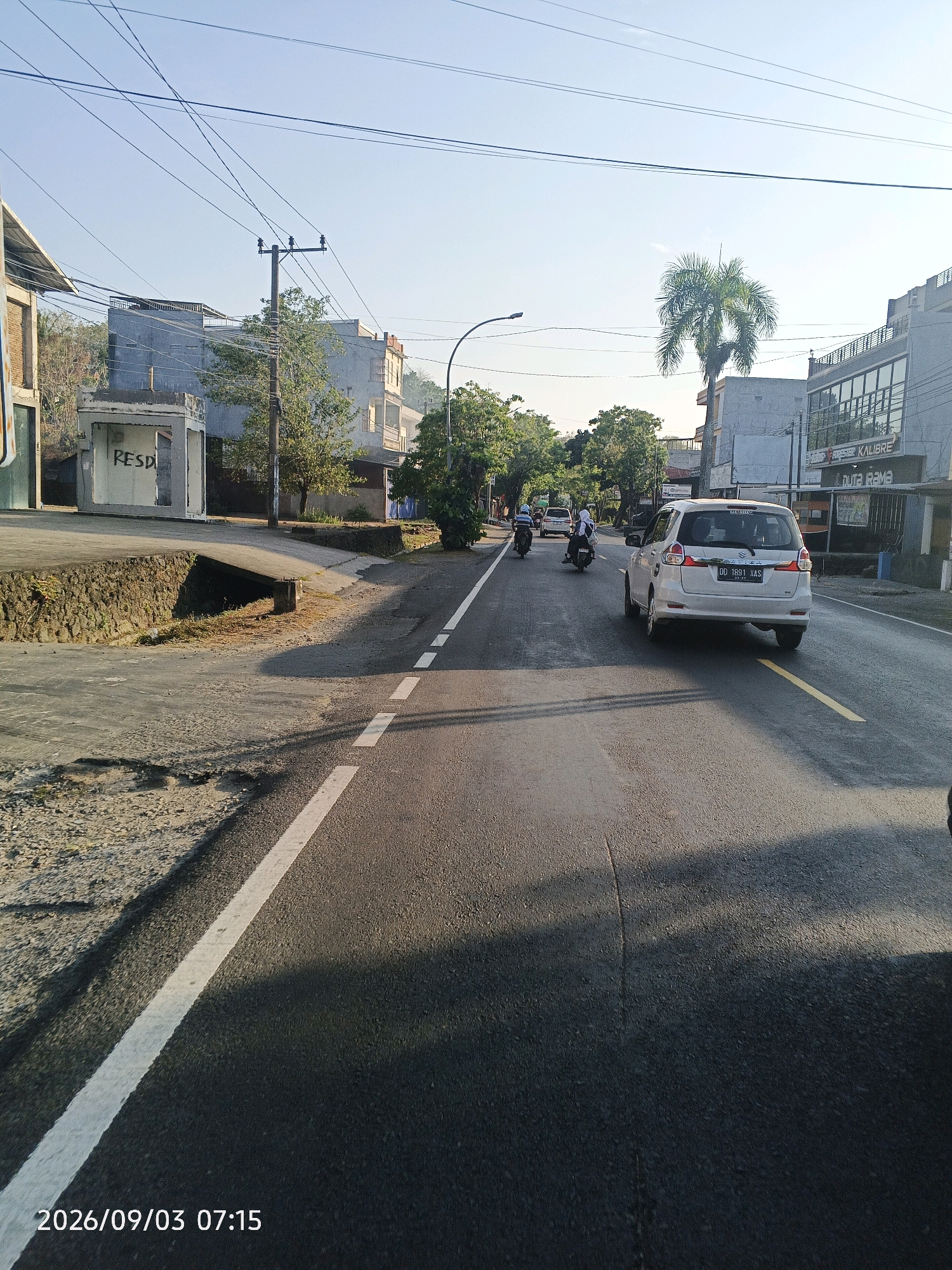 The image depicts a street scene in Pare-Pare, Indonesia, on a clear morning. The road is paved and has a white lane marking. In the foreground, the asphalt is visible, with shadows cast by trees. Several vehicles are present, including a white Suzuki Ertiga with license plate DD 1891 XAS, and two motorcycles, one with a rider wearing a white hijab. Buildings line the street, some appearing to be commercial establishments with signage. One building has a prominent "RESD" graffiti on its exterior. Overhead, power lines crisscross the sky. A palm tree stands tall in the background. The timestamp "2026/09/03 07:15" is visible in the bottom left corner of the image.