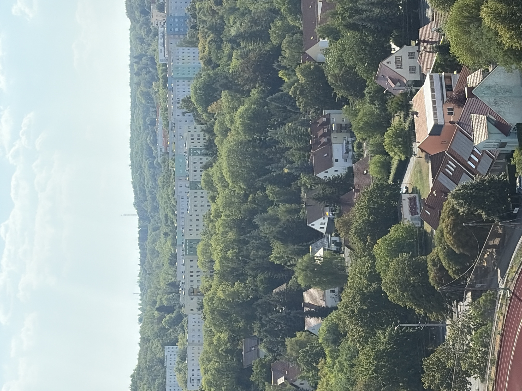 This image captures a dense urban and suburban landscape in Erfurt, Germany, characterized by a mix of residential buildings and lush greenery. The foreground features a section of a red athletic track, suggesting a sports facility. Beyond this, the scene transitions to a neighborhood of single-family homes with red-tiled roofs and some with solar panels, nestled amongst mature trees.
Further back, a line of multi-story apartment buildings, some painted in pastel shades of green, blue, and white, forms a stark contrast to the natural foliage. These apartment blocks appear to be characteristic of mid-20th-century Eastern European architecture. The buildings are situated on a gentle slope, with dense, mature trees occupying the spaces between and surrounding them, creating a verdant buffer.
In the distance, a hazy skyline is punctuated by a radio tower or similar transmission mast, indicating a broader urban or industrial area. The sky is a pale blue with scattered white clouds, suggesting a clear, possibly warm day. No people are clearly discernible in the image, and the overall impression is one of a well-established residential area integrated with significant amounts of natural vegetation. The lighting suggests it is daytime, likely late morning or early afternoon.