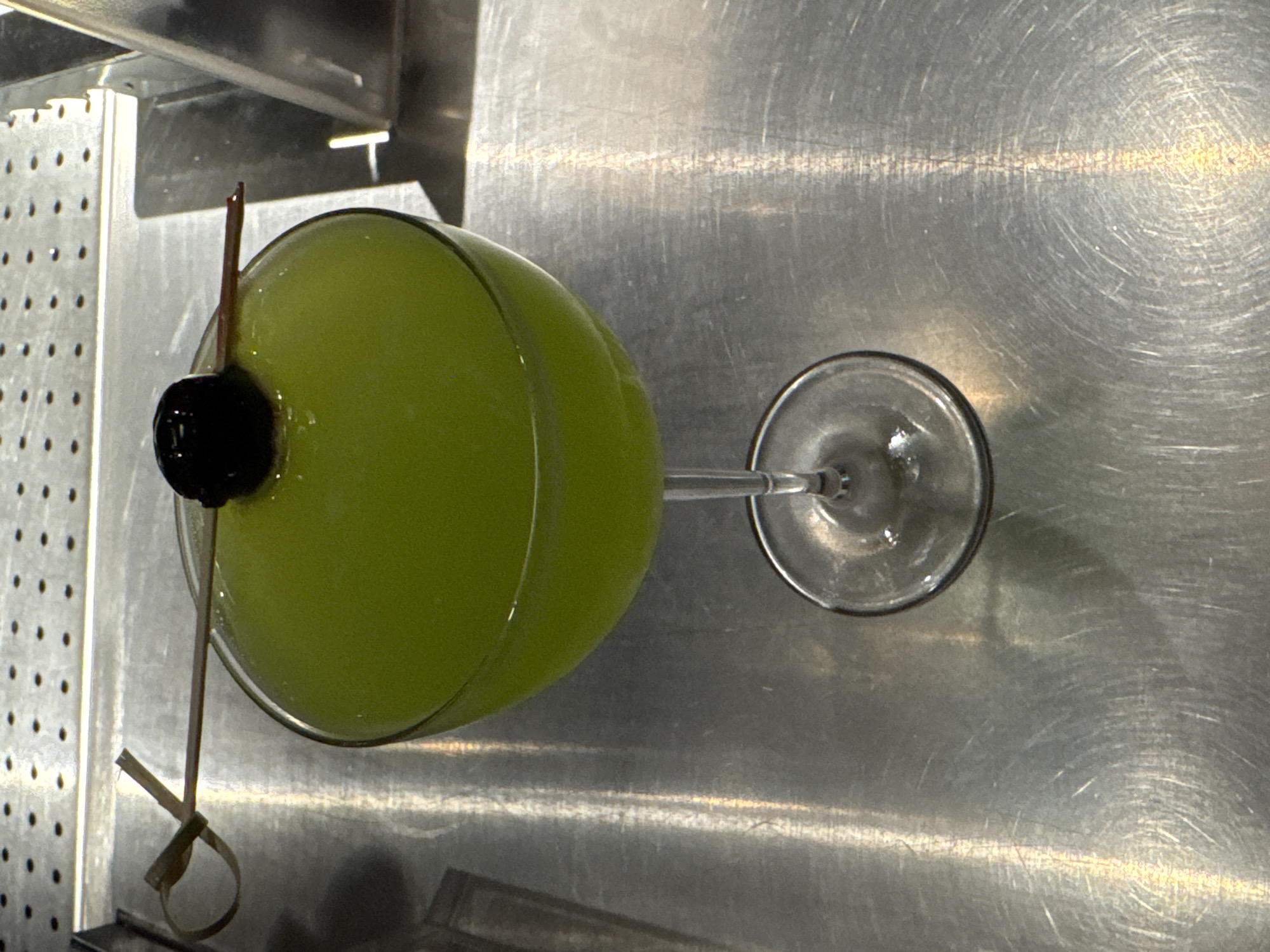 A green cocktail in a coupe glass, garnished with a cherry on a wooden skewer, is presented on a brushed metal surface. The skewer is secured to the rim of the glass with a twisted bamboo knot. The background features a perforated metal surface, likely part of a bar or kitchen setup. The lighting suggests an indoor environment with artificial illumination. The scene does not provide any information about its specific location in Yerevan, Armenia, or any individuals present.