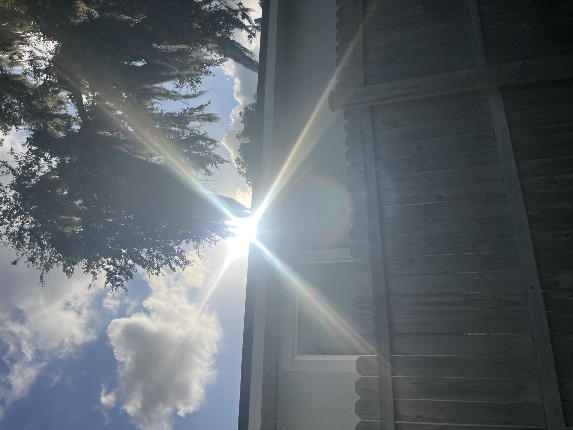 A sun flare obscures a view of a cloudy sky, the sun's rays emanating from the upper center of the frame and creating a rainbow effect. Below the sky, the top edge of a light-colored building is visible, with a dark-rimmed window. A wooden fence runs horizontally across the lower half of the image, with a tree branch, laden with Spanish moss, visible in the upper right corner. The image appears to have been taken outdoors on a sunny day.