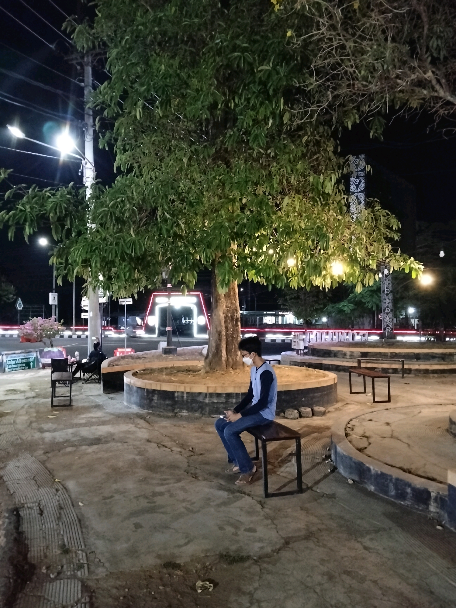 The image depicts an outdoor scene at night, possibly in Purworejo, Indonesia, centered around a large tree with decorative lights strung through its branches. A young man wearing a blue long-sleeved shirt, dark pants, and a face mask is seated on a dark metal bench. He is looking down, with his hands in his lap. The bench is placed on a circular concrete structure, which is surrounded by a low, dark-colored wall. The ground is paved with concrete and shows some cracks. In the background, there are streetlights, trees, and a large archway illuminated with neon lights. There are other benches and chairs scattered around the area. To the left of the main subject, another person is sitting in a foldable chair, partially obscured by the tree. There are also traffic signs and a small pink flowering bush visible in the distance. The overall atmosphere is dimly lit, with the artificial lights casting shadows.
