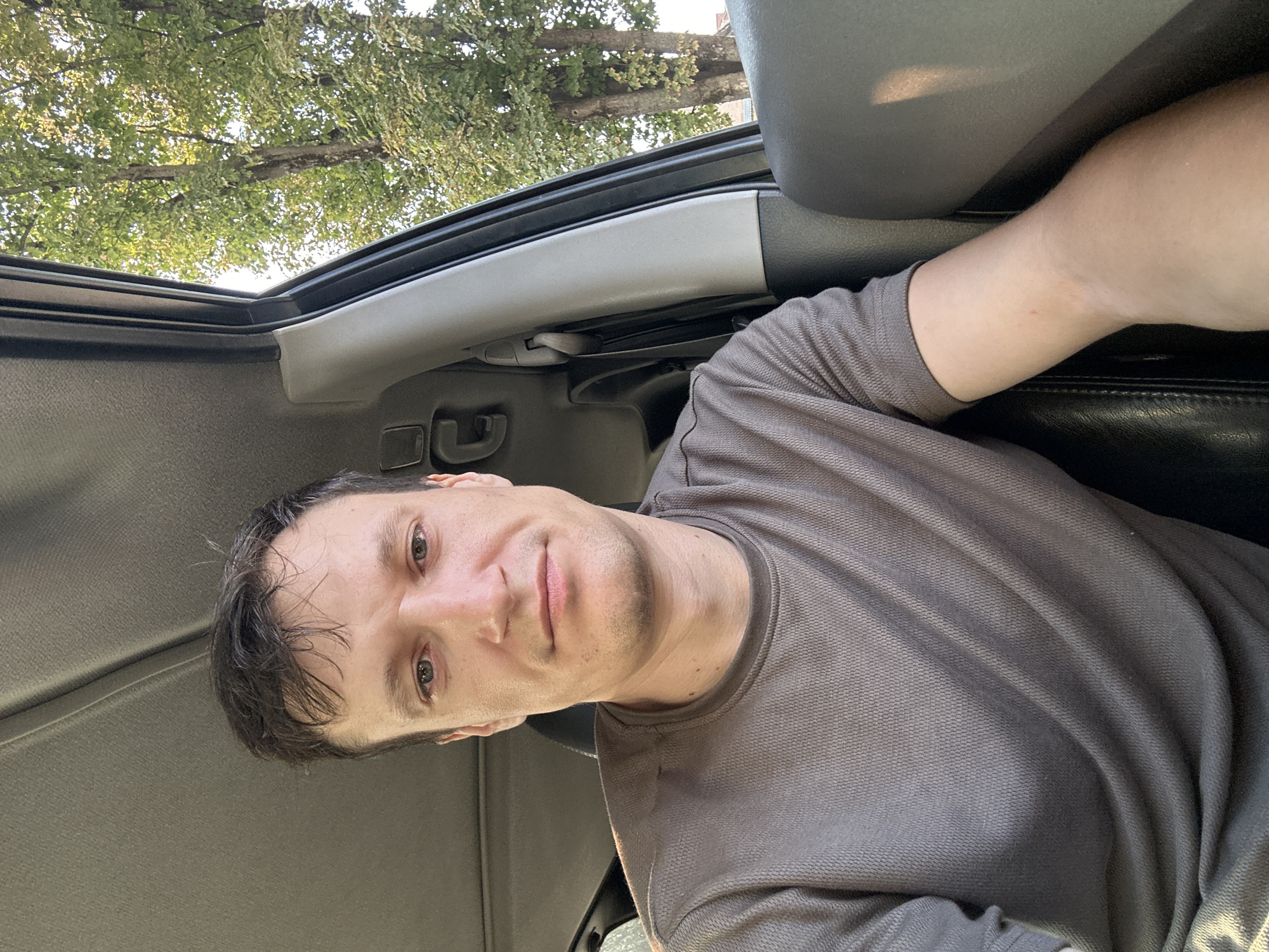 A man is captured from the chest up in a selfie taken from inside a vehicle. He is wearing a grey t-shirt and has short, dark hair. The background visible through the car window shows green trees and a bright sky, suggesting an outdoor daytime setting. The interior of the car, with its grey upholstery and black leather seat, is also visible. The location is Kamianets-Podilskyi, Ukraine.