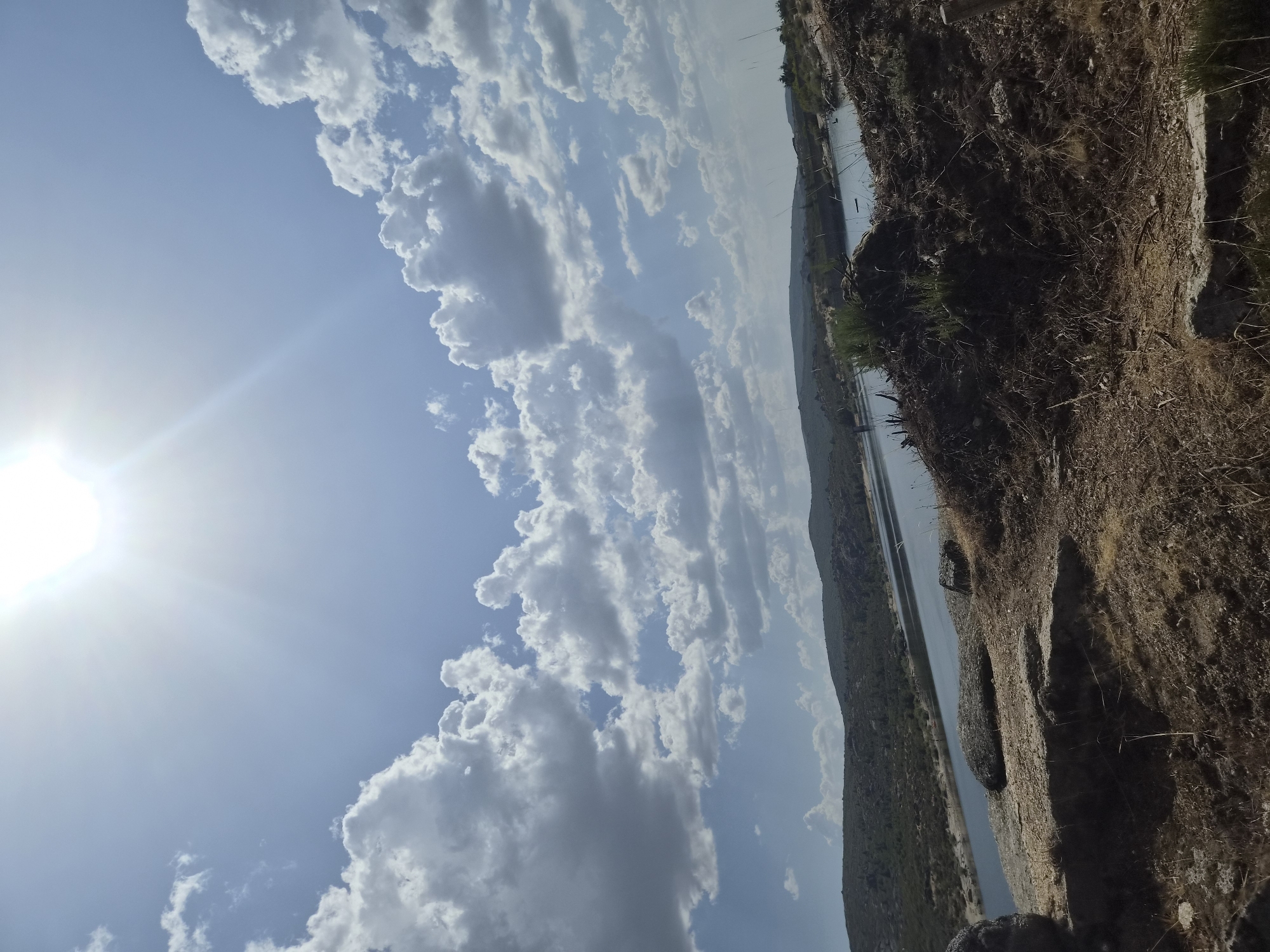 The image depicts a panoramic view of a reservoir in Gouveia, Portugal, under a bright, sunny sky with scattered clouds. The foreground features dry, rocky terrain with sparse vegetation, including dried grasses and small shrubs. Several large rocks are present in the immediate foreground. The middle ground consists of the calm surface of the reservoir, reflecting the sky and surrounding landscape. The far side of the reservoir is lined with rolling hills covered in dense green vegetation, suggesting a natural, possibly mountainous, environment. The sun is high in the sky, casting a bright light and creating a slight lens flare in the upper portion of the image. There are no people or animals visible in the scene. The overall atmosphere is one of natural beauty and tranquility.