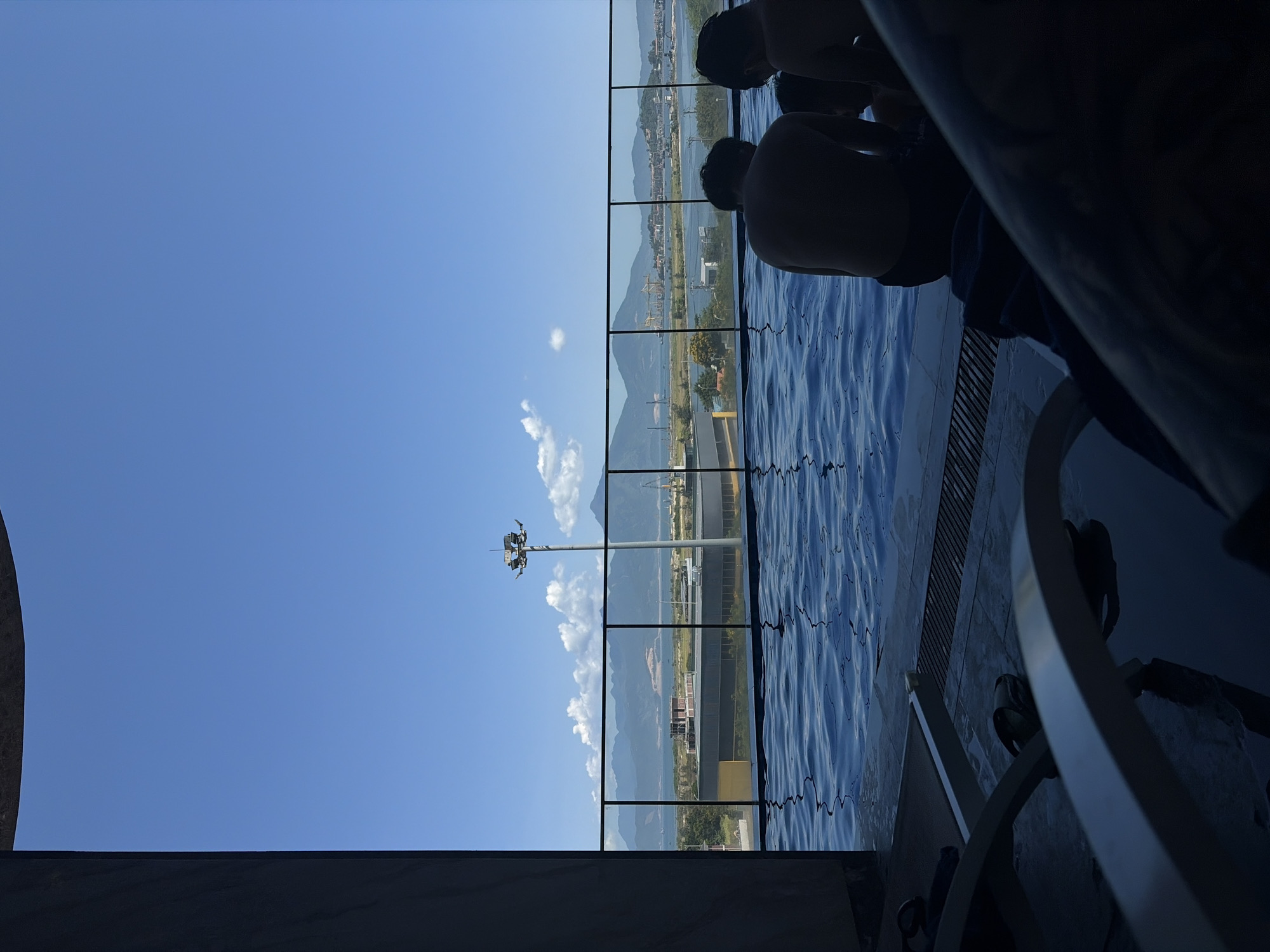 The image depicts a panoramic outdoor scene captured from inside what appears to be an infinity pool or a pool featuring large glass barriers. In the immediate foreground, on the right side, the backs of two individuals are visible, partially submerged in the pool's rippling blue water. They are silhouetted against the bright background, seemingly observing the expansive view.
Large, clear glass panels, demarcated by dark vertical frames, separate the pool area from the outside landscape. Beyond these panels, a vast expanse of bright blue sky, adorned with scattered white clouds, dominates the upper portion of the scene. Distant mountains form a majestic backdrop across the horizon, stretching through the middle ground. Below the mountains, a body of water, likely a river or bay, can be seen, with various urban and industrial structures dotting its shores. A tall light pole, equipped with multiple fixtures, stands prominently outside the glass barriers on the left side of the frame.
The setting suggests a modern resort or hotel with a scenic overlook, consistent with the coastal and mountainous geography of Da Nang, Vietnam. The activity taking place is leisure and relaxation in the pool, enjoying the view. The time of day is bright daylight, likely midday, with clear, sunny weather. The scene evokes a feeling of tranquility and enjoyment of nature. There is no visible text in the image.