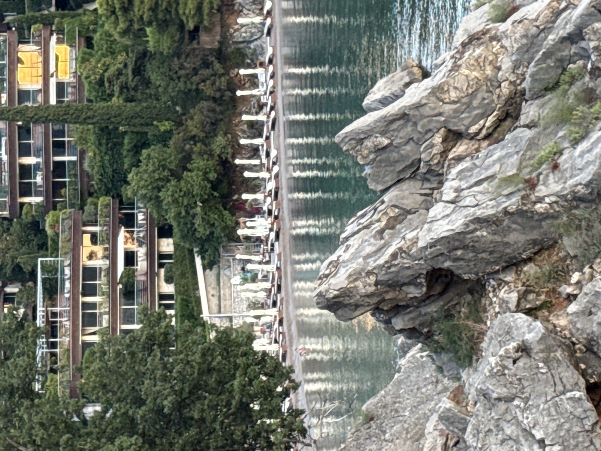 This image depicts a coastal scene likely in Budva, Montenegro, focusing on a rocky foreground leading to a beach with loungers and umbrellas, and a hotel building situated on a hillside in the background. The foreground is dominated by large, grey, textured rocks, some with sparse green vegetation growing between them. Beyond the rocks, a sandy beach is visible, lined with numerous white beach umbrellas and empty loungers. Two individuals are in the shallow water near the shore, appearing to be swimming or wading. The sea is a calm, shimmering turquoise with reflections of the umbrellas and the sky. Behind the beach, a multi-story hotel building is built into a lush green hillside, featuring balconies and large windows, some of which are illuminated with a warm, golden light, suggesting sunset or interior lighting. Tall, slender cypress trees are interspersed with the vegetation around the hotel. The overall impression is a serene coastal resort setting.