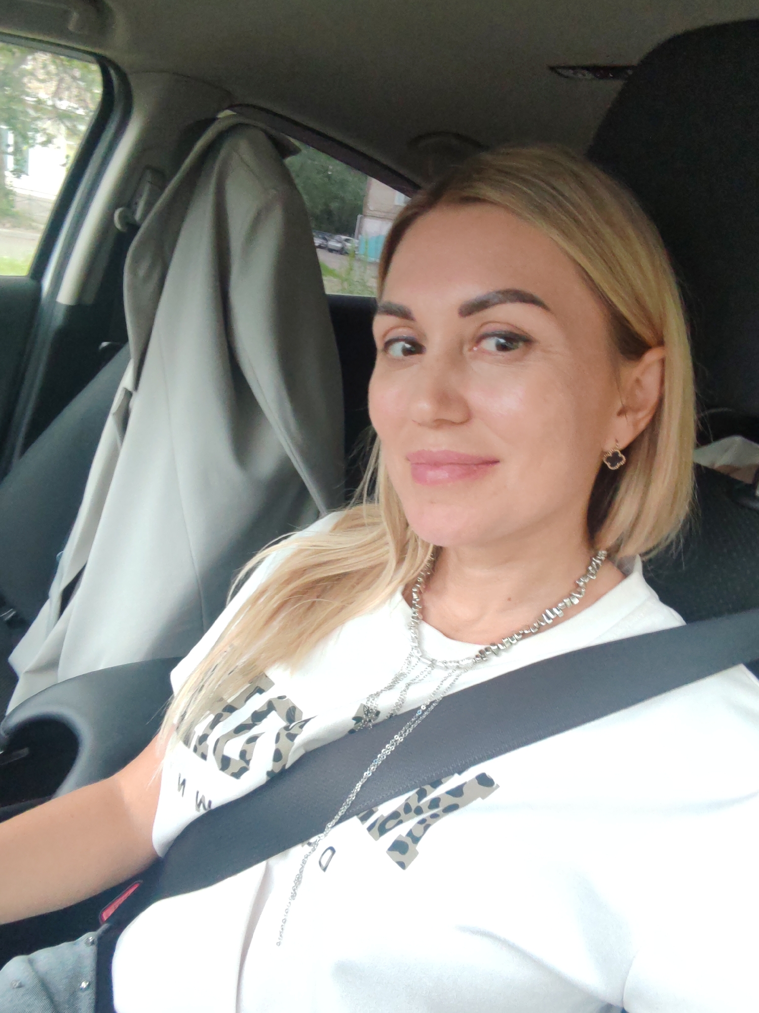 In a selfie taken from inside a car, a woman with blonde hair smiles as she looks towards the camera. She is wearing a white t-shirt with a leopard print graphic and silver necklaces. A black seatbelt is visible across her chest. A light-colored jacket is hanging on the seat behind her. The background shows a blurred view of trees and buildings through the car windows, suggesting an outdoor setting during the daytime. The woman's expression appears relaxed and happy. Visible text on her t-shirt includes "W M" in black lettering. The context indicates the location is Komsomolsk-on-Amur, Russia.