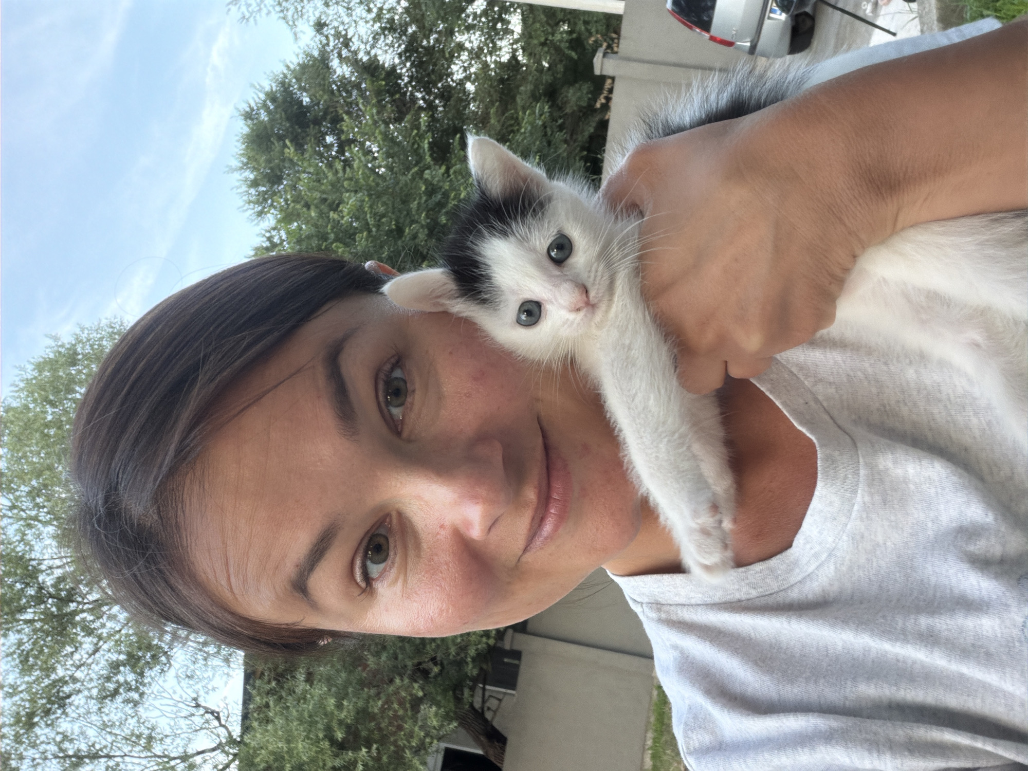 This close-up portrait, oriented sideways, features a woman gently holding a small kitten outdoors. The woman, with dark hair and light brown/hazel eyes, looks towards the camera with a soft, subtle smile. She wears a light-colored (possibly grey) t-shirt.
Cradled securely in her arm and resting near her shoulder is a young kitten, predominantly white with black patches on its head, ears, and back. The kitten has piercing blue eyes and appears calm and alert, gazing directly forward.
The background indicates an outdoor setting during daytime, with ample natural light. Lush green trees are visible, suggesting a natural or semi-rural environment, consistent with Podlastva (Poljice), Montenegro. Portions of a building and the rear of a silver car are also discernible in the upper right background. No text is visible in the image. The scene conveys a moment of affection and tenderness between the woman and the kitten.