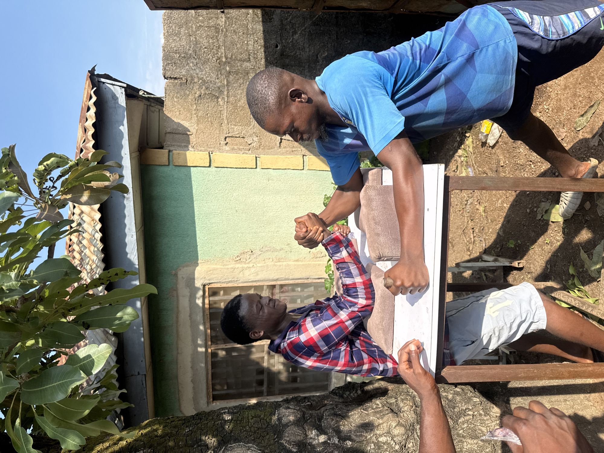 Two young African men are engaged in an arm-wrestling match outdoors. They are positioned opposite each other at a makeshift table. The man on the left, wearing a red, blue, and white plaid shirt and light-colored shorts, is leaning forward, gripping the other man's hand. The man on the right, dressed in a blue t-shirt with a geometric pattern and dark shorts, is also leaning in, their hands locked in a struggle. Their faces show concentration and exertion. The setting appears to be a residential area in Akwanga, Nigeria, with a building with a green facade, a corrugated metal roof, and a brick wall in the background. Sunlight casts shadows on the ground, indicating it is daytime. A tree trunk is visible on the left side of the frame.