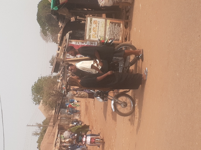 This image depicts a bustling street scene in Gwadanye, Nigeria, under bright, possibly hazy, daylight conditions. The ground is an unpaved, reddish-brown dirt surface.
In the mid-ground, three individuals are clustered around a silver and black motorcycle. A female, positioned to the left, wears a long-sleeved black top, a long black skirt or dress, and white flip-flops; her light-colored hair is long and flows over her shoulders. She faces toward the right. A male in a light-colored short-sleeved shirt and dark trousers is seated on the motorcycle, facing left, towards the female. To the right of the motorcycle, another male stands, wearing a black short-sleeved top, dark shorts, and dark flip-flops; he also faces left, engaging with the other two individuals. The motorcycle has chrome front wheel spokes and handlebars.
To the immediate right, a wooden sign with a dark frame advertises "THOMCY GLOBAL ENTERPRISE P.O.S SERVICES AVAILABLE," followed by a list of financial services including "Account Opening," "Buy Enrollment," "Talk Deposit," "Cash Withdrawal," "Fund Transfer," and "Bill Payment."
The background reveals a row of market stalls constructed with corrugated metal roofs and wooden supports, stretching along the left side of the street. Multiple pedestrians and individuals on motorcycles are visible, some carrying goods. Power lines are discernible overhead. Distant structures, including one with a pyramidal roof, and scattered trees complete the background.