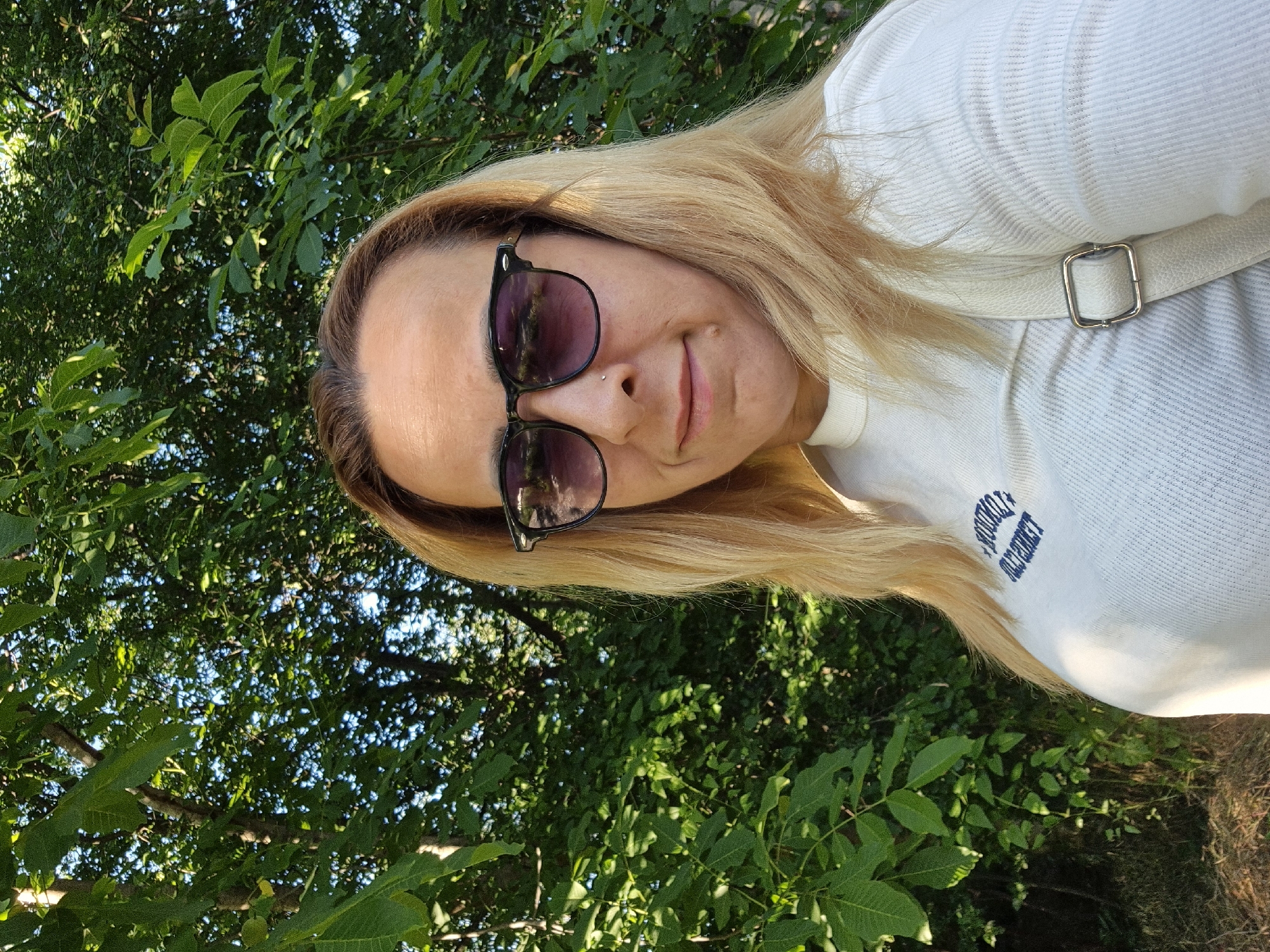 A medium shot, taken outdoors on a sunny day, shows a woman with blonde hair and sunglasses posing for a selfie. She is wearing a white ribbed t-shirt with blue text on it and a light-colored strap that goes over her shoulder. The background is filled with lush green trees and dappled sunlight. The woman is looking directly at the camera with a slight smile. She has a nose ring.