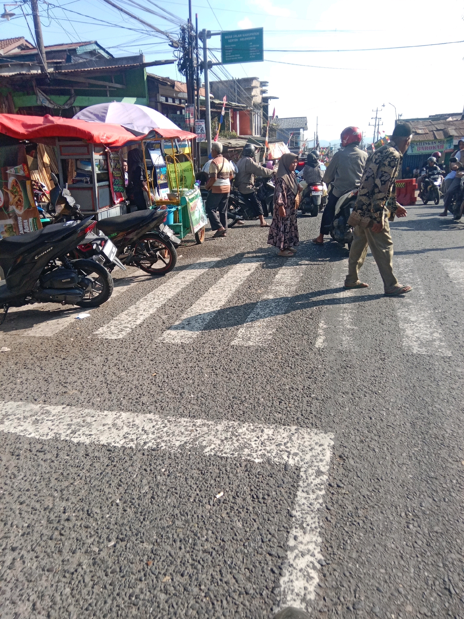 The image depicts a street scene in Kertek, Indonesia, characterized by a pedestrian crossing and surrounding activity. In the foreground, a zebra crossing is visible on an asphalt road, with white markings. To the left, two motorcycles are parked, one black and the other partially visible. Behind the motorcycles, a food stall with a red awning and a purple umbrella is set up, displaying various items and signage.
Several individuals are present in the scene. A man in a camouflage patterned shirt and tan pants is walking across the zebra crossing, his shadow cast on the road. Another person, wearing a red helmet, is seated on a motorcycle to the right of the pedestrian. A woman in a patterned dress and hijab is also visible in the middle ground, near the food stall, and another woman in a black hijab stands behind the stall. Further in the background, more people are seen near motorcycles and stalls, suggesting a busy market or roadside area. Buildings with traditional Indonesian architecture line the street, and overhead, a complex network of electrical wires is suspended. A green directional signpost with text in Indonesian is also mounted on a pole. The overall environment is outdoors, under a bright, clear sky, indicating daytime.