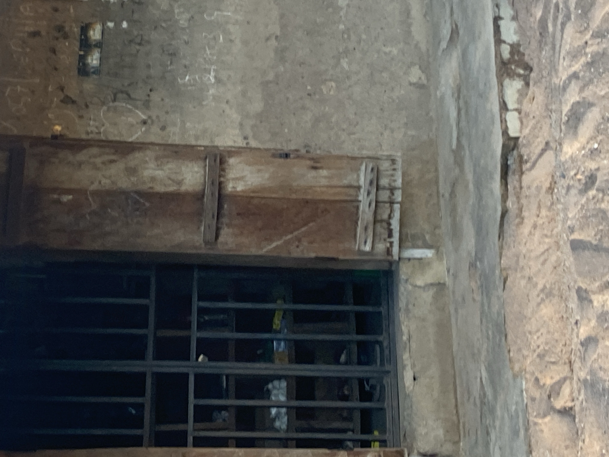 The image shows the exterior of a building in Jauro Yinu, Nigeria. To the left, a barred opening, likely a window or door, reveals shelves inside stocked with various items. The bars are dark, and the interior is dimly lit. To the right of the barred opening is a wooden shutter, partially closed, secured with horizontal planks. The wall of the building is rough and textured, with visible graffiti and markings, including what appears to be a heart shape and some illegible writing. The ground in front of the building is uneven, with a concrete ledge and sandy areas. The overall impression is one of a weathered and lived-in structure.