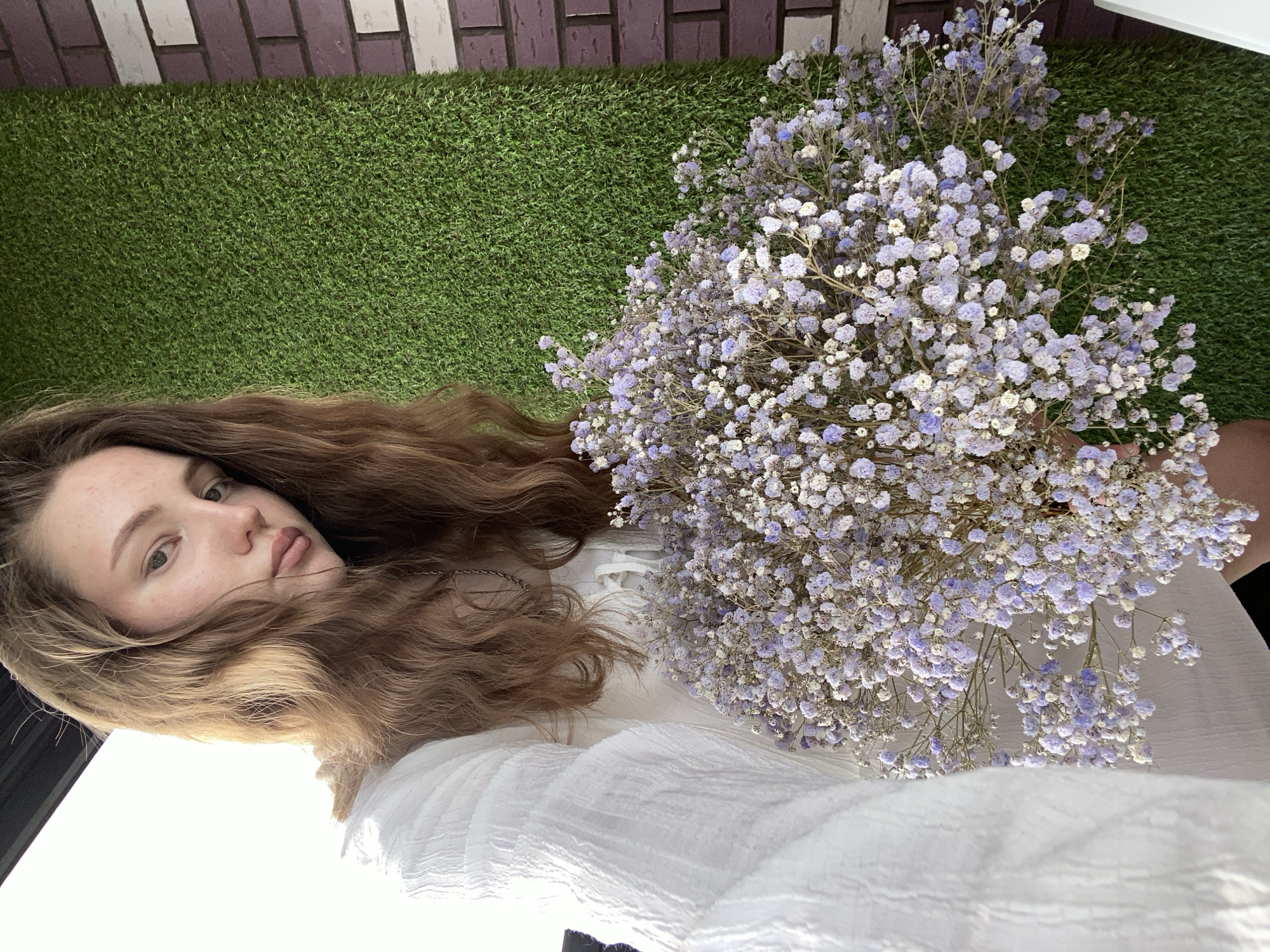 A young woman with long brown hair lies on artificial green turf, next to a large bouquet of purple and white baby's breath flowers. She is wearing a white, loose-fitting top. The background includes a wall resembling gray and white brick. The location is Izhevsk, Russia.