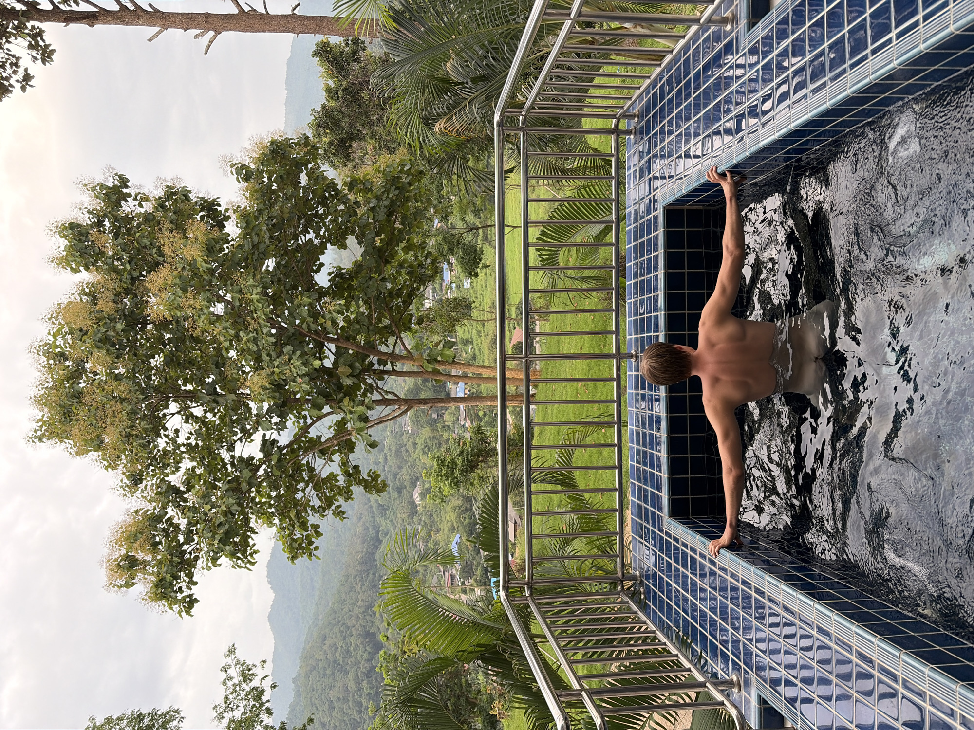 A man with blond hair is submerged in a dark blue tiled hot tub. The hot tub is outdoors, on a balcony or patio, overlooking a lush green landscape. The man is facing away from the camera, with his arms resting on the edge of the hot tub. The background consists of rolling hills covered in dense vegetation, with a few scattered buildings visible in the distance. Tall trees, including palm trees, frame the scene. The sky is overcast. The location is identified as Ban Kao Dua, Thailand.