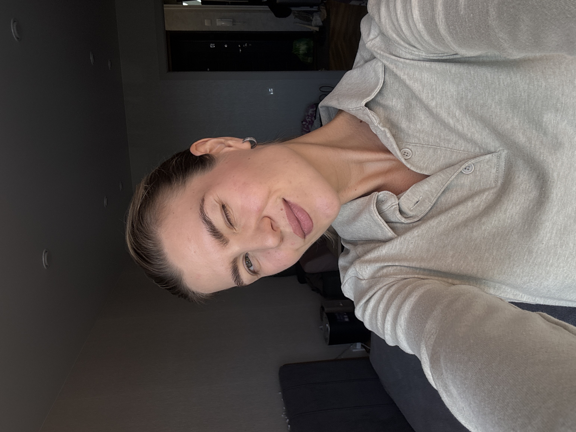 A rotated indoor selfie captures a young woman from the chest up, looking playfully towards the camera. Her head is tilted to her right, and she exhibits a mischievous expression with one eye winked or partially closed and her tongue slightly poking out. She has light brown hair, pulled back, and natural-looking makeup with defined eyebrows and a subtle pinkish-mauve lip color. A silver-colored hoop earring is visible in her right ear.
She is wearing a light grey or off-white long-sleeved top made of a textured, possibly heathered fabric. The top features a collar and a placket with two visible buttons at the neckline.
The background is dimly lit, revealing a domestic interior with dark walls. On the right, part of a dark sofa or chair is visible, along with what appear to be shelves or electronic devices further back. The subdued lighting creates an intimate and casual atmosphere. There is no visible text in the image.