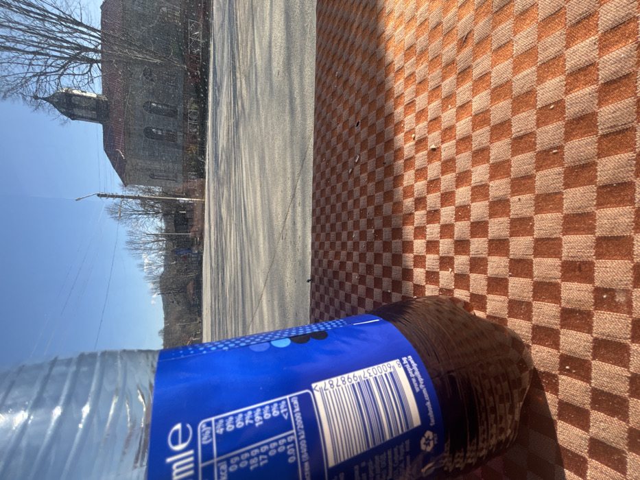 The scene is set in Brezovo, Bulgaria, under clear blue skies with direct sunlight. The foreground features a partially visible clear plastic bottle of Pepsi, angled slightly, with its dark brown liquid content and blue label prominently displayed. The label shows nutritional information in white text, a barcode, and Bulgarian text indicating "pepsi.bg" and "facebook.com/PepsiBulgaria". The bottle rests on a brown and light brown checkered textile surface, likely a tablecloth, exhibiting minor debris.
Beyond the immediate foreground, a wide, empty expanse of light grey asphalt pavement, resembling a public square or parking area, is visible. Long, distinct shadows are cast across this paved surface from an unseen source, indicating a low sun angle. In the mid-ground, a tall, grey utility pole with overhead power lines stands.
The background is dominated by a historical-style stone building with a red tiled roof and a multi-tiered bell tower, consistent with a church. The building features arched windows. To the right and behind the building, several large deciduous trees with bare branches are present. Distant hills or mountains can be seen on the horizon. There are no individuals present in the visible area.