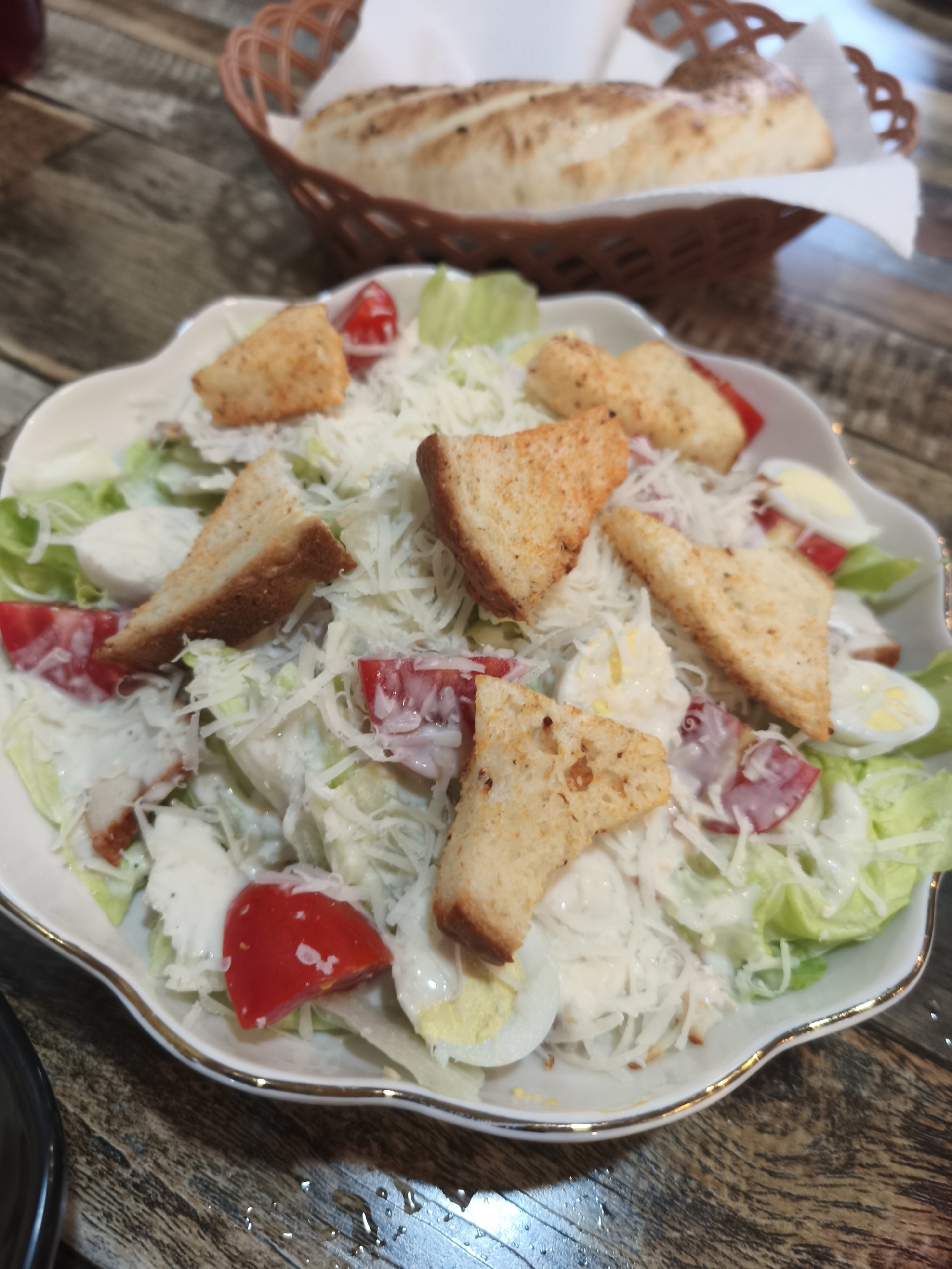 This image shows a close-up of a Caesar salad served in a white, fluted bowl with a gold rim. The salad contains chopped romaine lettuce, croutons, cherry tomato halves, hard-boiled egg slices, shredded Parmesan cheese, and visible pieces of grilled chicken. The salad is dressed with a creamy white dressing. In the background, out of focus, is a woven brown basket containing two pieces of bread. The salad is placed on a wooden table with water droplets on its surface, suggesting it may have been recently prepared or served. There are no people or other discernible objects in the immediate vicinity of the salad. The setting appears to be a restaurant, though no specific identifying features are present to pinpoint the location within Asan-si, South Korea.