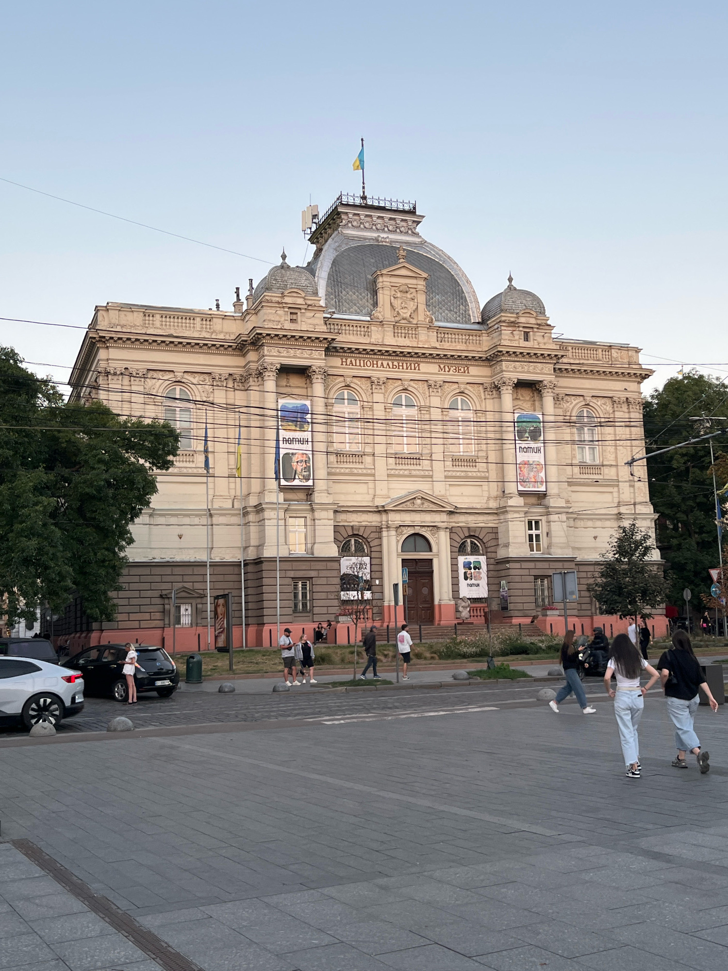 The image shows the National Museum in Lviv, Ukraine, from its exterior. The museum is a large, ornate building with a prominent dome and neoclassical architectural elements. The facade is light-colored, with decorative carvings and large arched windows. Two Ukrainian flags are visible on poles in front of the building, and banners with posters are hung on the facade. A Ukrainian flag flies from the top of the dome.
In the foreground, a paved plaza area is visible, with several people walking and standing. The area is bordered by a street with cars parked along the side and a road with a few vehicles, including a white car and a black electric car. A few trees and greenery are also present in the scene, with a dense green tree on the left side of the image. The sky is clear and light blue, indicating it is daytime. The overall impression is of a public space in front of a significant cultural institution.
