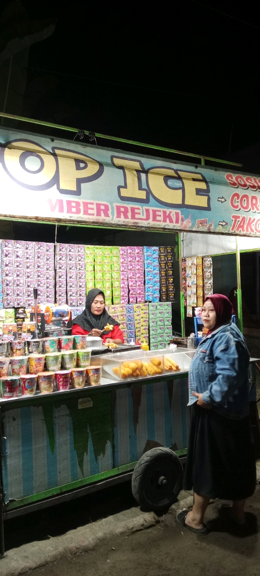 A nighttime street scene in Kesamben, Indonesia, features a food stall with a bright green awning. The stall displays "TOP ICE" in large yellow letters. Beneath this, pink text reads "MBER REJEKI." Further to the right, red text indicates "SOSI" and "COR," and white text reads "TAKO." The stall is lined with colorful rows of packaged goods, predominantly in shades of pink, green, blue, and black, with some resembling cup noodles or snacks.
Two women are visible. One woman, wearing a black hijab and a red top, stands behind the stall, preparing food. She is holding a skewer with small, light-colored food items over a cooking surface. In front of her are various stacked cups. The second woman, wearing a maroon hijab and a denim jacket over a dark skirt, stands to the right of the stall, facing away from the camera. She appears to be a customer or an associate.
In the foreground, a clear plastic display case holds a collection of fried foods, likely pastries or snacks. To the left of the stall, a black tire from a vehicle is visible. The ground is paved with rough material, and the overall lighting suggests nighttime conditions.
