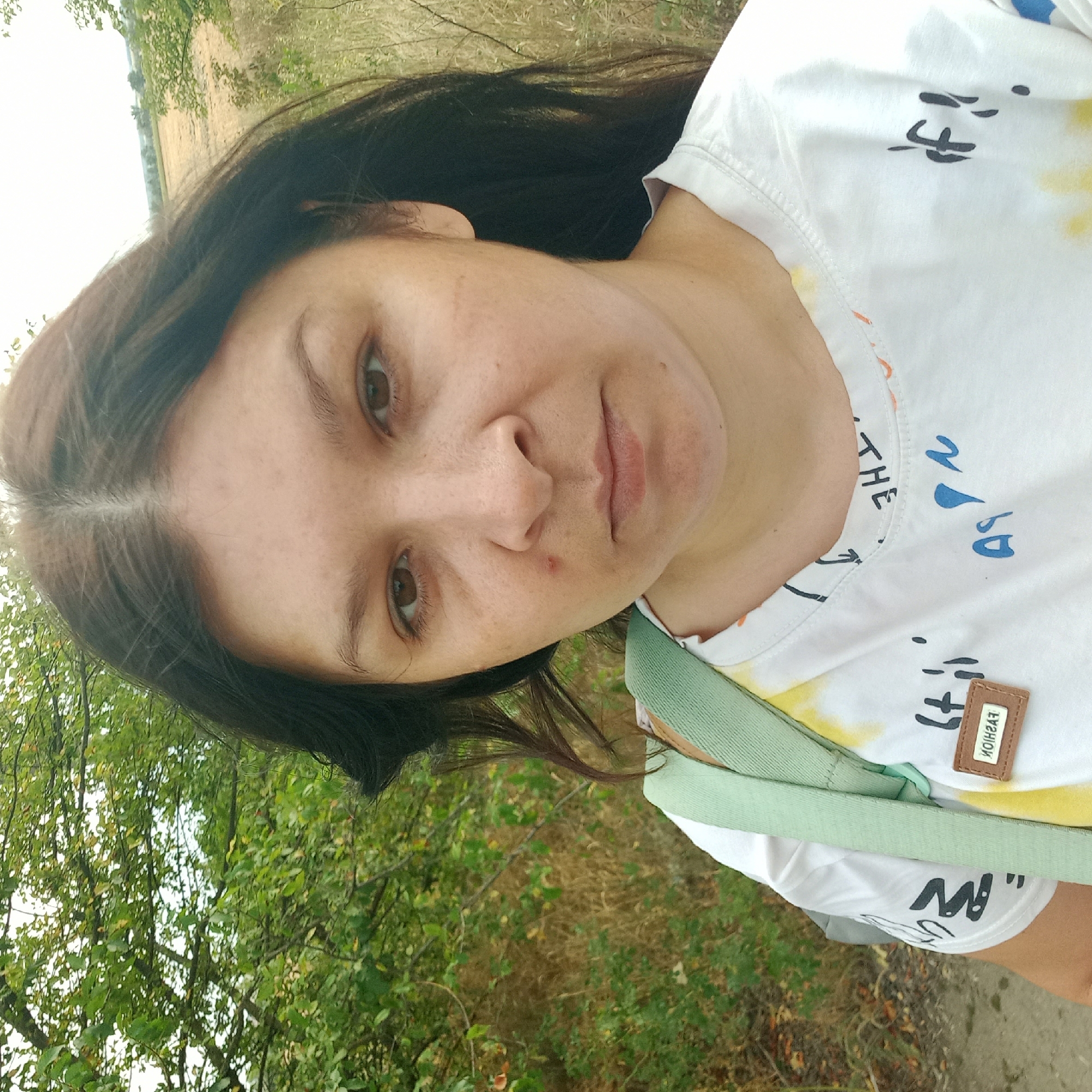 A woman with dark hair and brown eyes is captured in a close-up, outdoor selfie. She is wearing a white t-shirt with a colorful, abstract design and a small brown tag with the word "FASHION" in reverse. A light green strap of a bag is visible over her left shoulder. She has a small reddish mark on her cheek. The background consists of lush green trees and dry grass, with a hint of a field and trees in the distance, suggesting a rural or semi-rural setting. This photograph was taken in Neopalymivka, Ukraine.