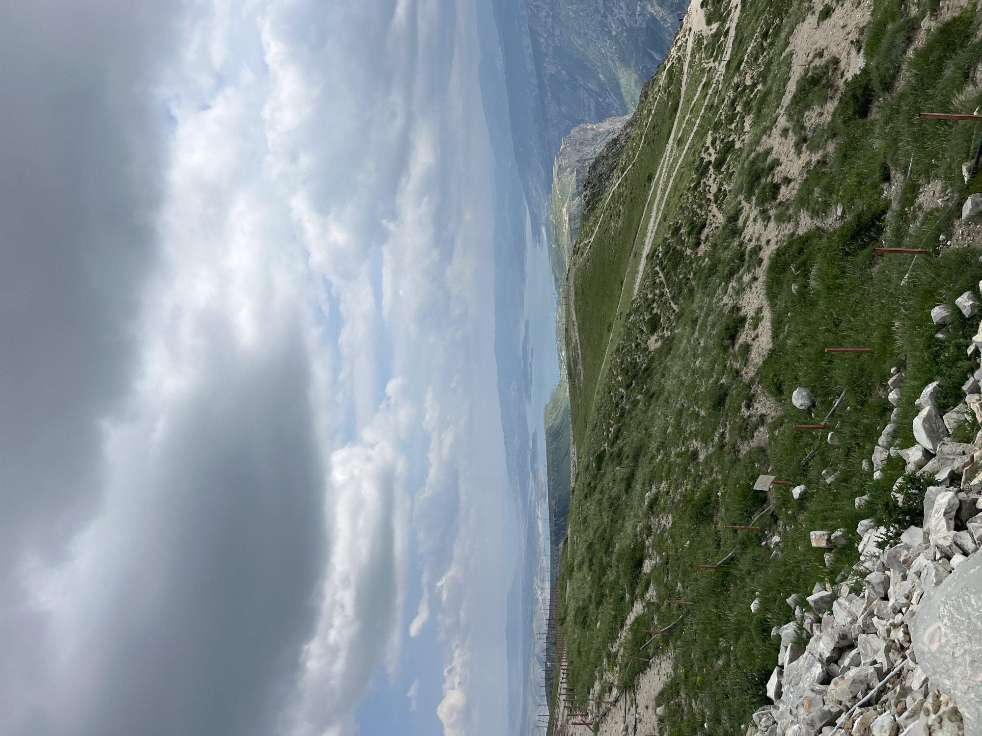 The image shows a view from a high vantage point, likely a mountain or steep hillside, overlooking a body of water and distant mountains. The foreground consists of rocky terrain transitioning into a grassy slope, with several small fence posts visible. A winding path can be seen traversing the hillside. The sky is cloudy, with a mix of light and dark cloud formations. The body of water stretches into the distance, separating the near hillside from the mountains on the opposite shore. The overall scene suggests a natural landscape setting. There is no evidence of people present in this image. Chirkey, Russia, is not discernable from this photograph.