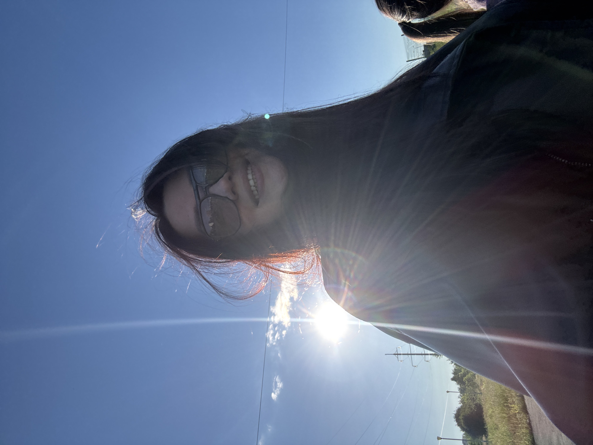 The image captures a selfie taken outdoors on a bright, sunny day. The main subject is a person with long dark hair, seen from a low angle, wearing dark sunglasses and smiling broadly, revealing their teeth. A significant sun flare emanating from the lower right of the frame indicates direct sunlight, casting strong light rays across the image. The sky is a clear, vibrant blue with a few sparse, wispy clouds. Faint, thin lines, likely power lines or wires, stretch horizontally across the sky in the upper portion of the image. In the background, towards the lower right, there is natural green foliage, including trees and grass, alongside what appears to be a road or path edge and a power pole structure. A small portion of another person's dark hair is visible in the top right corner of the frame. The subject's smile suggests a cheerful or happy demeanor. No visible text is present in the image.