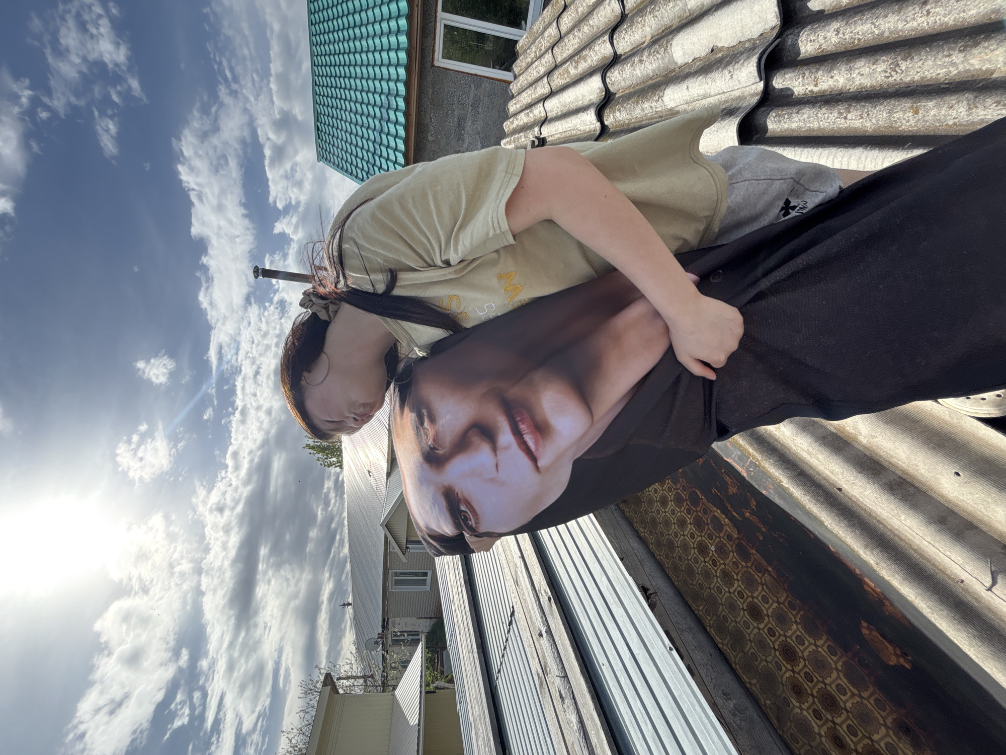 A person is on a roof, holding a pillow with a face printed on it. The background shows residential buildings, suggesting a suburban area. The sky is partly cloudy. The location is near Фоки, Russia.