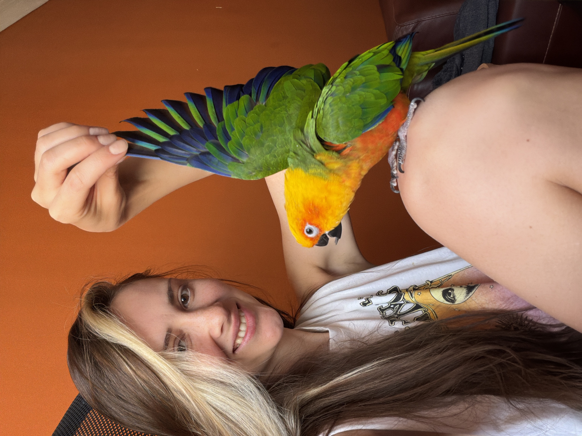 A woman with long brown and blonde hair is holding a colorful parrot. The parrot is mostly green, yellow and orange, with blue and black wing feathers. The woman is smiling and wearing a white t-shirt with a graphic design. The background is a plain orange wall, and a brown leather object. The location and information about Khimki, Russia, is not identifiable within the content of the image.
