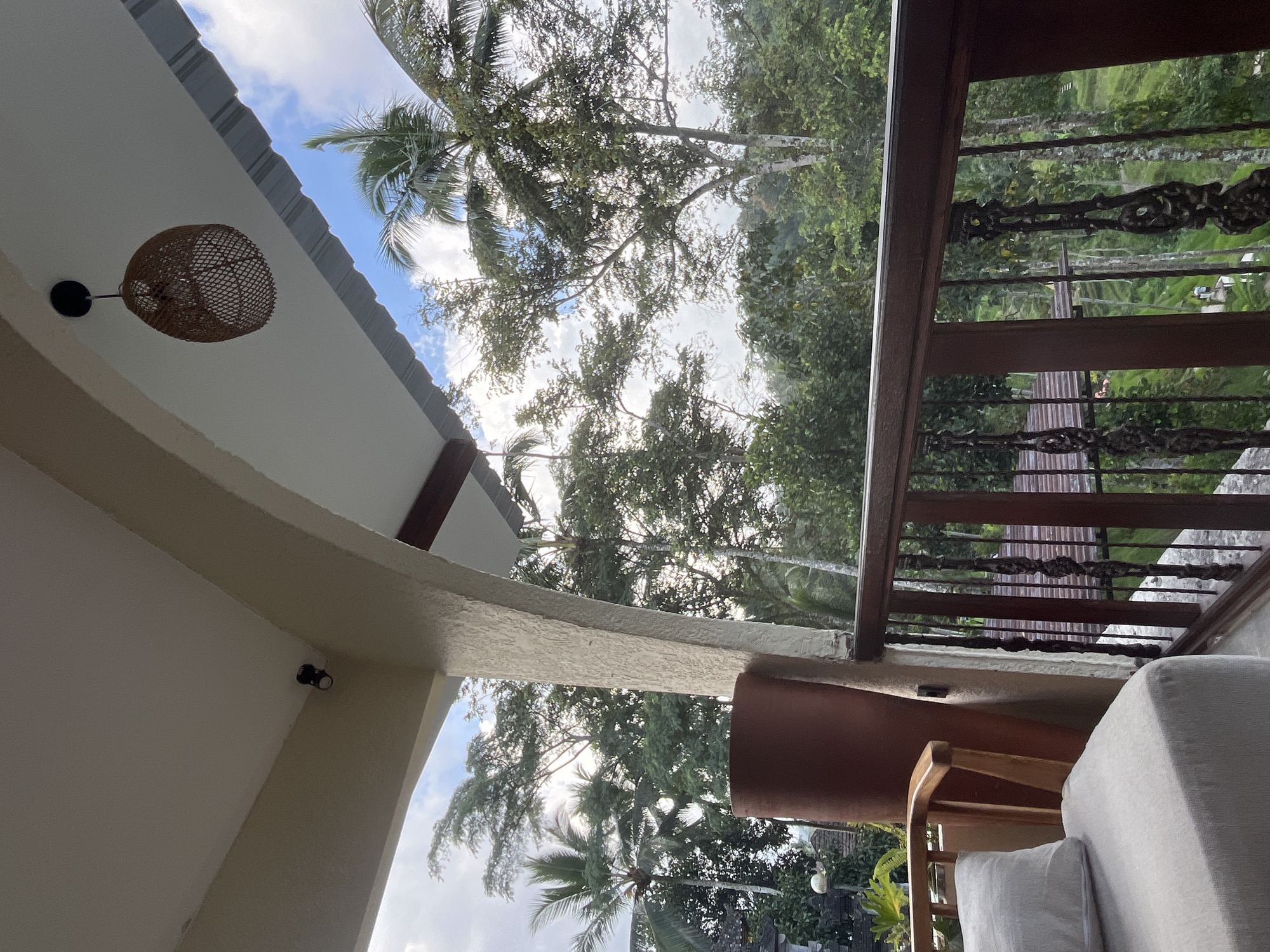 The image, taken from a covered outdoor space like a balcony or terrace, offers a serene view of a lush tropical environment. The perspective suggests the original image was rotated 90 degrees clockwise.
In the immediate foreground, a light-colored, possibly stuccoed, ceiling or overhang is visible, from which a round, woven rattan light fixture hangs. A small, dark security camera is mounted on an archway of the structure. To the right, a sturdy wooden balcony railing features decorative, dark metal scrollwork inserts. Part of a light-grey upholstered outdoor sofa or cushion and the back of a wooden chair are also visible on the balcony floor, suggesting a comfortable lounging area.
Beyond the balcony, the background is filled with dense, vibrant green foliage of various trees, including tall palm trees, indicative of a tropical climate. Bright sunlight filters through the leaves, casting a soft glow. The sky above is a clear blue with scattered white clouds, suggesting a pleasant daytime, possibly late morning or early afternoon. The overall atmosphere is peaceful and natural, characteristic of a retreat setting.
The location context indicates this scene is in Tegallalang, Indonesia. There is no visible text in the image.
