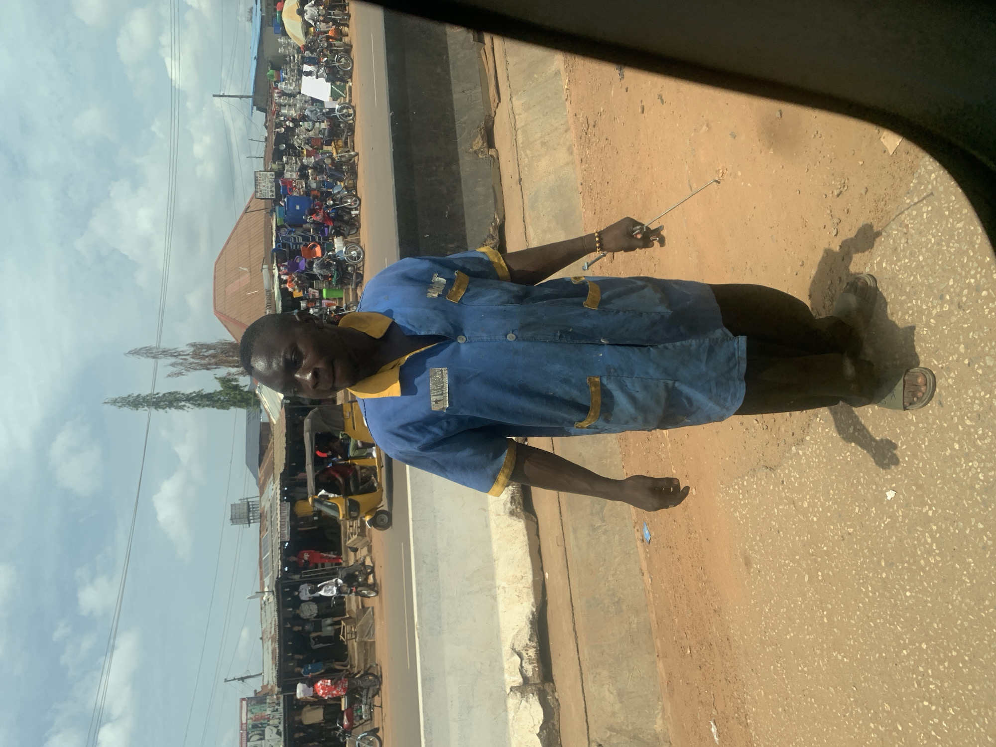 A street scene in Ado, Nigeria, photographed from inside a vehicle. An African man wearing a blue shirt with yellow trim stands near the roadside, facing the camera. He is holding a thin metal object in his right hand. A busy commercial area is visible across the road, with shops and a large number of motorcycles and auto-rickshaws (Keke Napep) parked along the street. The sky is partly cloudy. The road surface is a mix of concrete and dirt. The man is wearing sandals.