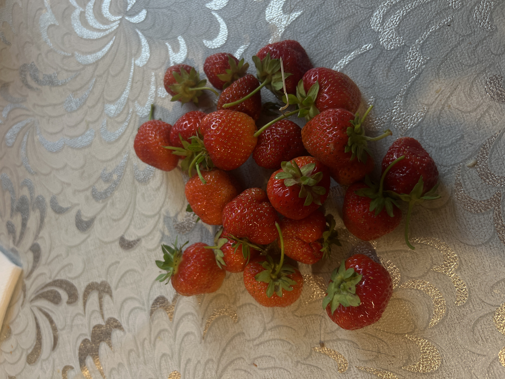 A pile of ripe red strawberries is scattered on a textured, light-colored tablecloth with a swirling, decorative pattern in shades of silver and gold. The strawberries vary slightly in size and shape, with some showing their green leafy tops. The lighting appears to be natural or from an overhead source, casting subtle shadows on the tablecloth. The image does not contain any information about the city of Афанасьево, Russia.