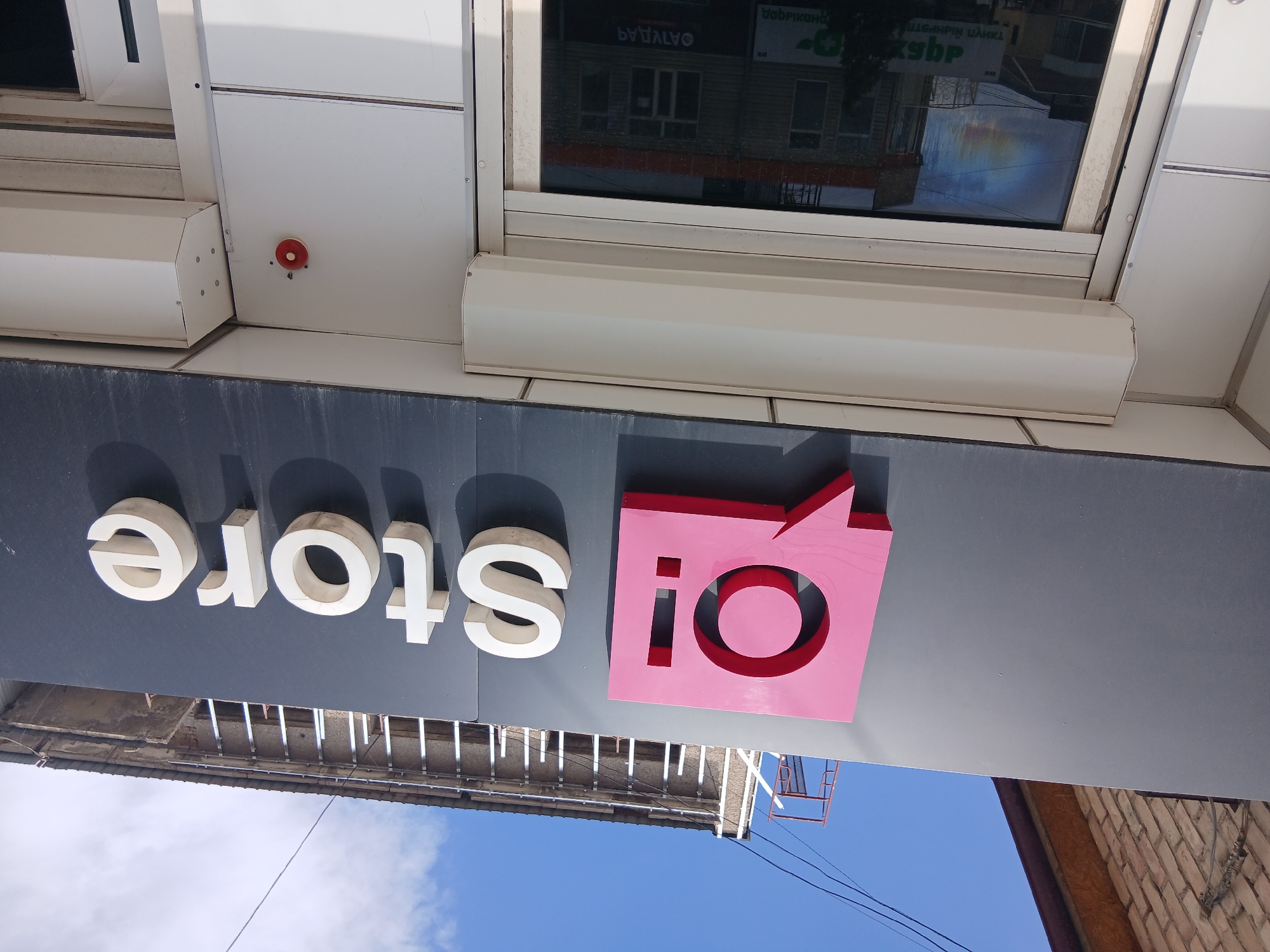The image shows the exterior of a store with a prominent sign. The sign features a pink speech bubble logo with a "q!" inside, followed by the white text "Store". The store has a dark gray facade with white lettering. Below the sign, there is a closed white roller shutter over a large window. A small red circular alarm bell is mounted on the white facade to the right of the roller shutter. In the reflection on the window, buildings and some signs with Cyrillic text are visible. Above the store, the facade of an adjacent building with brickwork and scaffolding is visible against a blue sky with some clouds. The image is taken from a low angle, looking upwards towards the store sign and the building above. This storefront is located in Talas City, Kyrgyzstan.